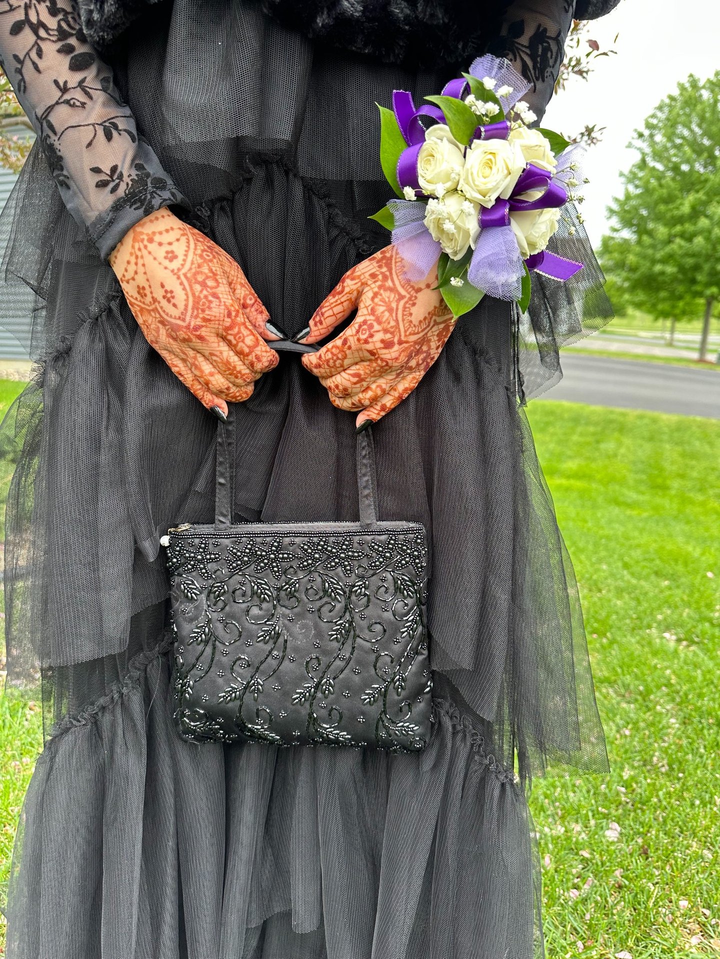 Close up of two hands with henna on them, holding a purse strap, wearing a black dress & corsage