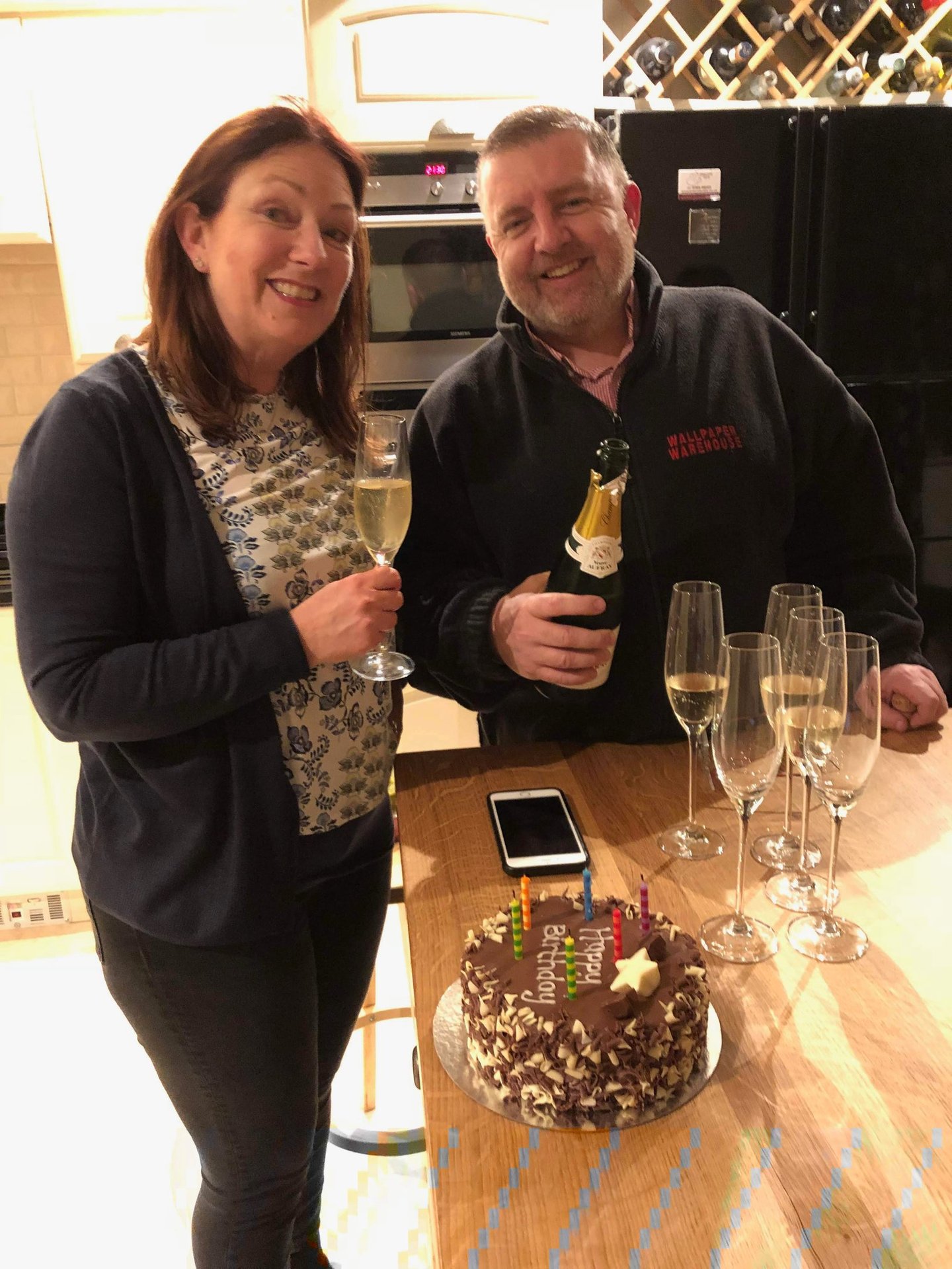 Two adults celebrating birthday in modern kitchen with champagne and cake reading 'Happy Birthday