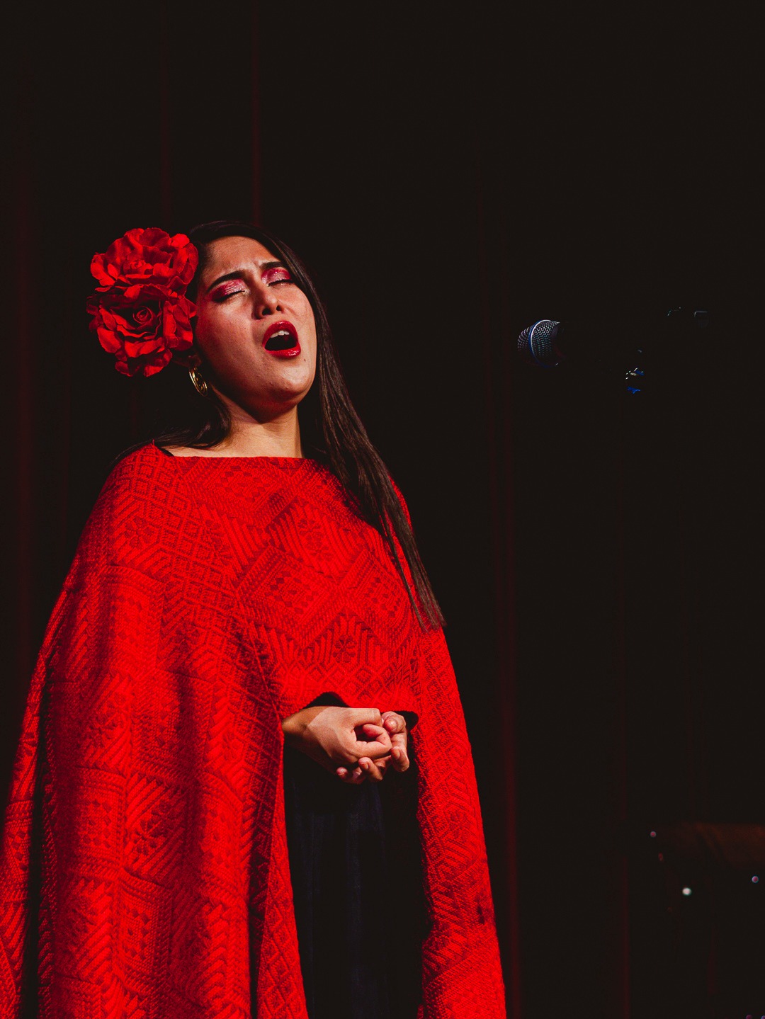 Female singer performing in a red embroidered poncho with a flower in her hair at a live concert.