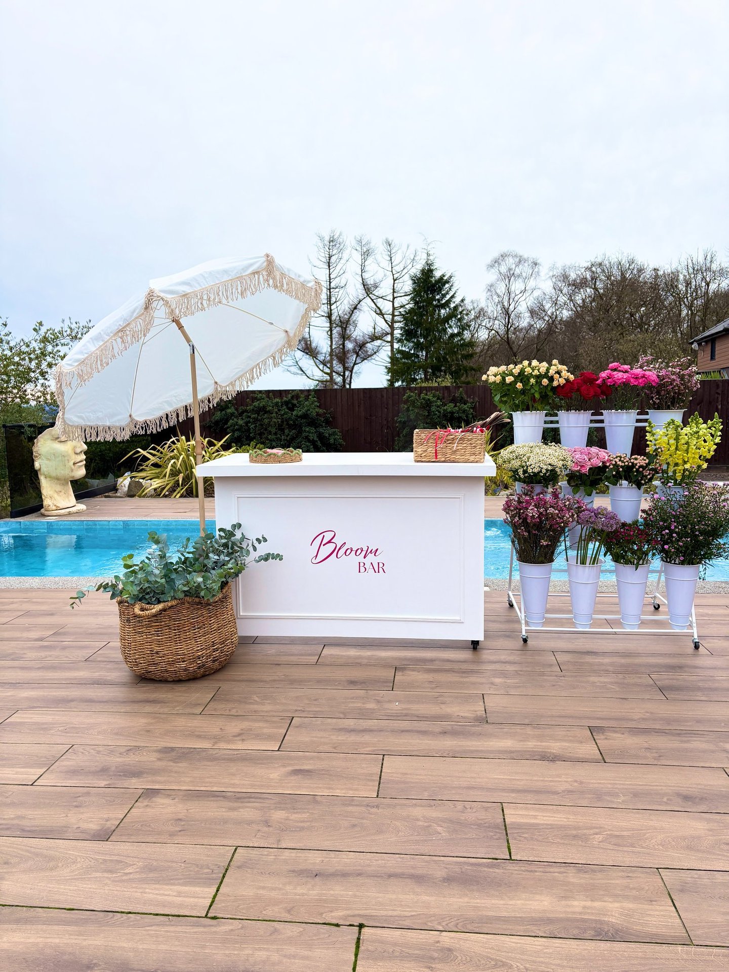 Mobile bloom bar flower cart with white fringed umbrella and fresh floral arrangements by a poolside deck.