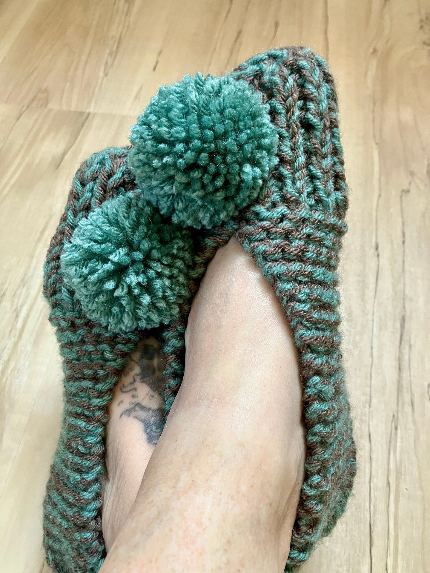 close-up of sage green and taupe colored hand knit slippers on models crossed feet