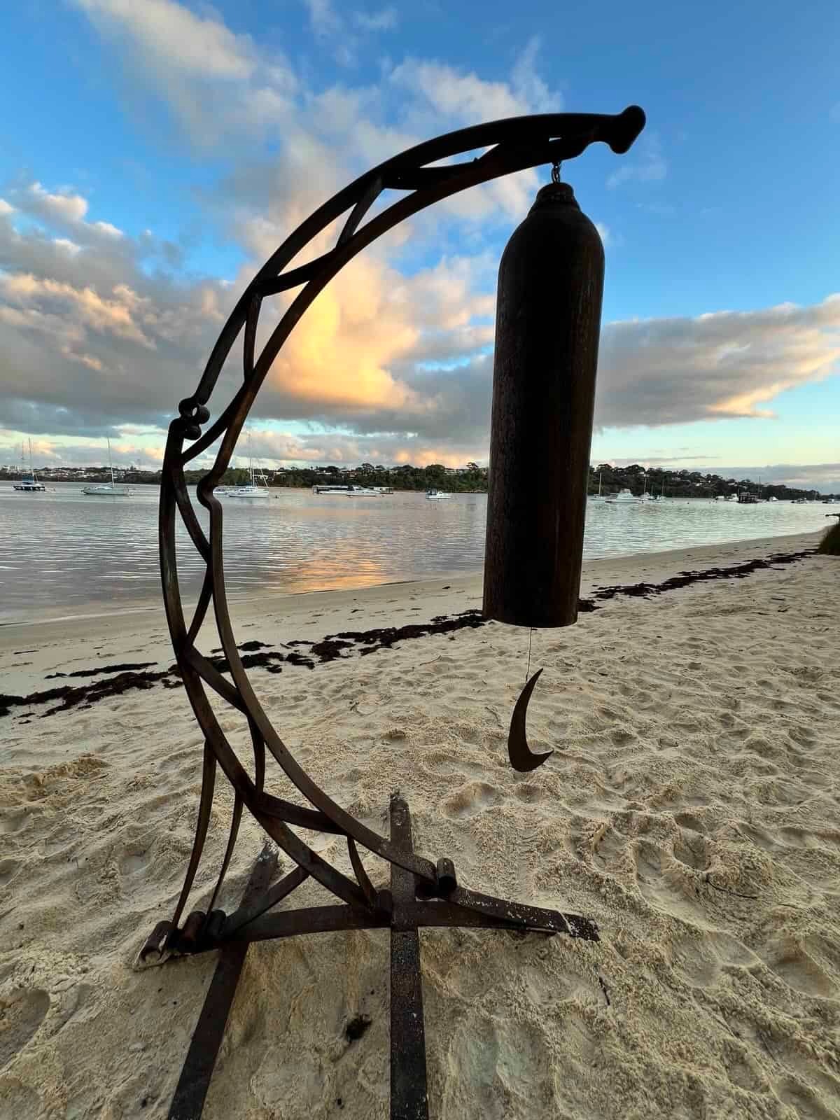 Rustic metal bell sculpture on a sandy beach at sunset with boats in the coastal bay.