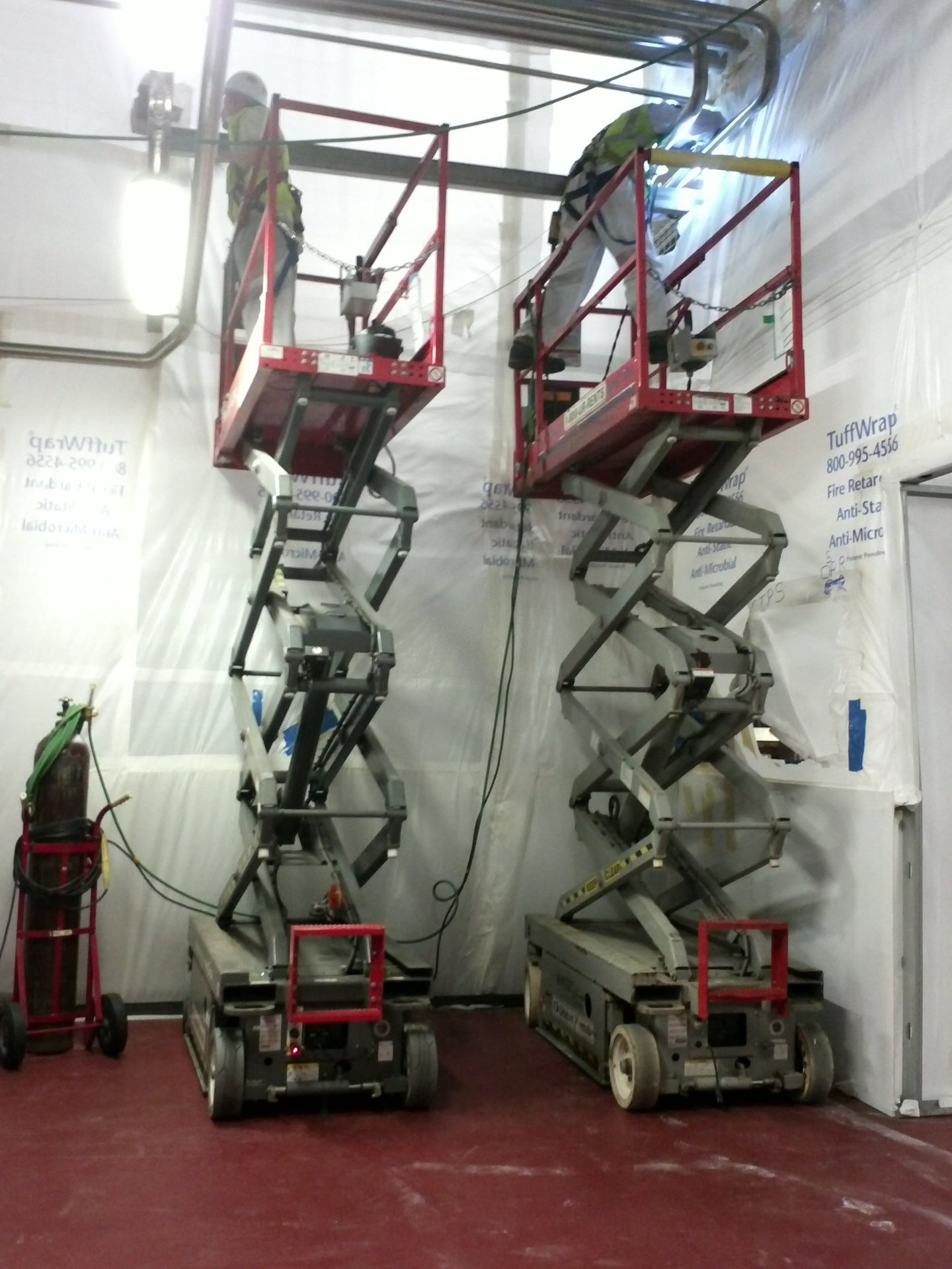 Two construction workers on scissor lifts performing pipe installation in dust containment area.