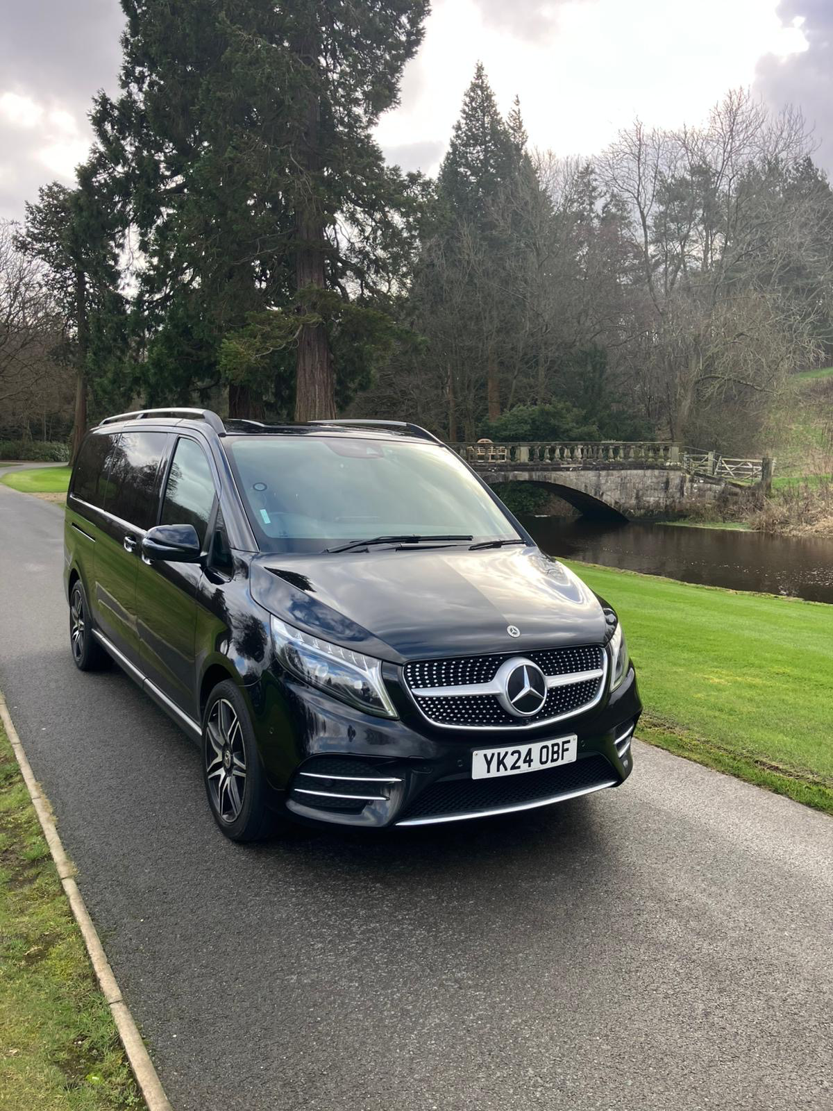 Black Mercedes-Benz V-Class luxury MPV parked on a scenic road near a stone bridge and river.