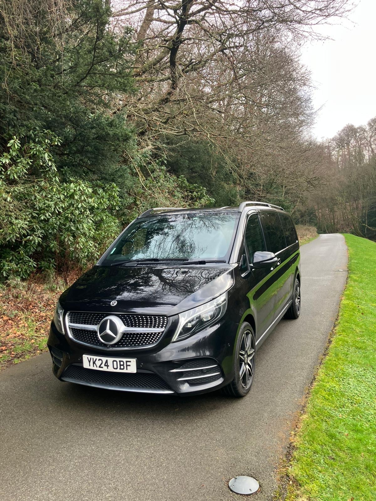 Black Mercedes-Benz V-Class luxury passenger car on a scenic rural road near woodland.