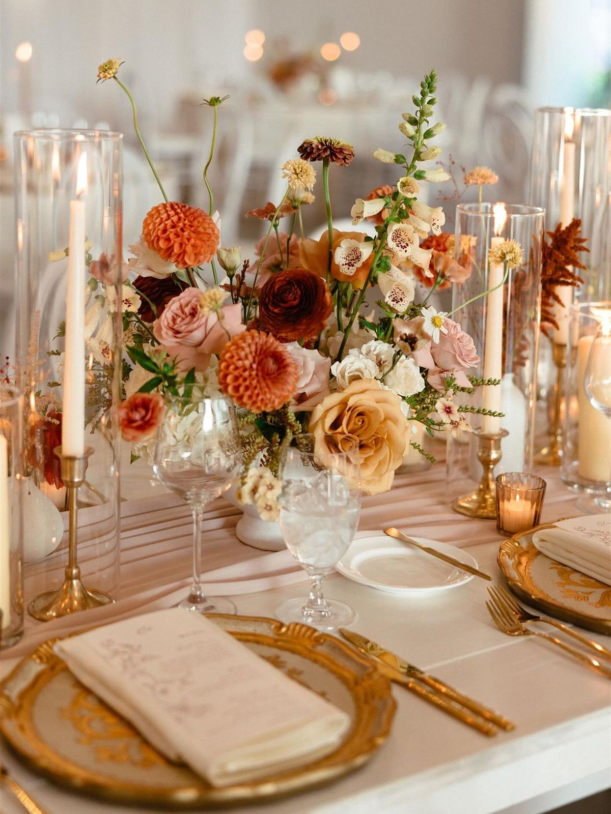Mesa elegante con vajilla dorada, velas altas y arreglo floral sofisticado.