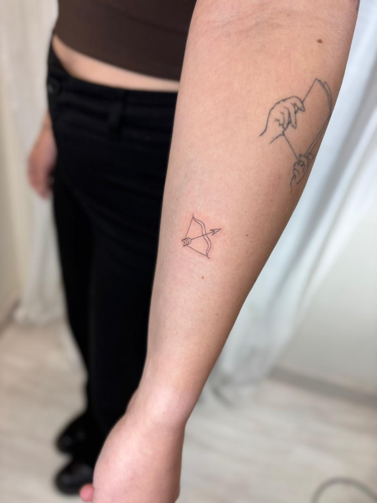 Tatuaje fineline en Madrid de arco y flecha en trazo continuo y minimalista en el antebrazo