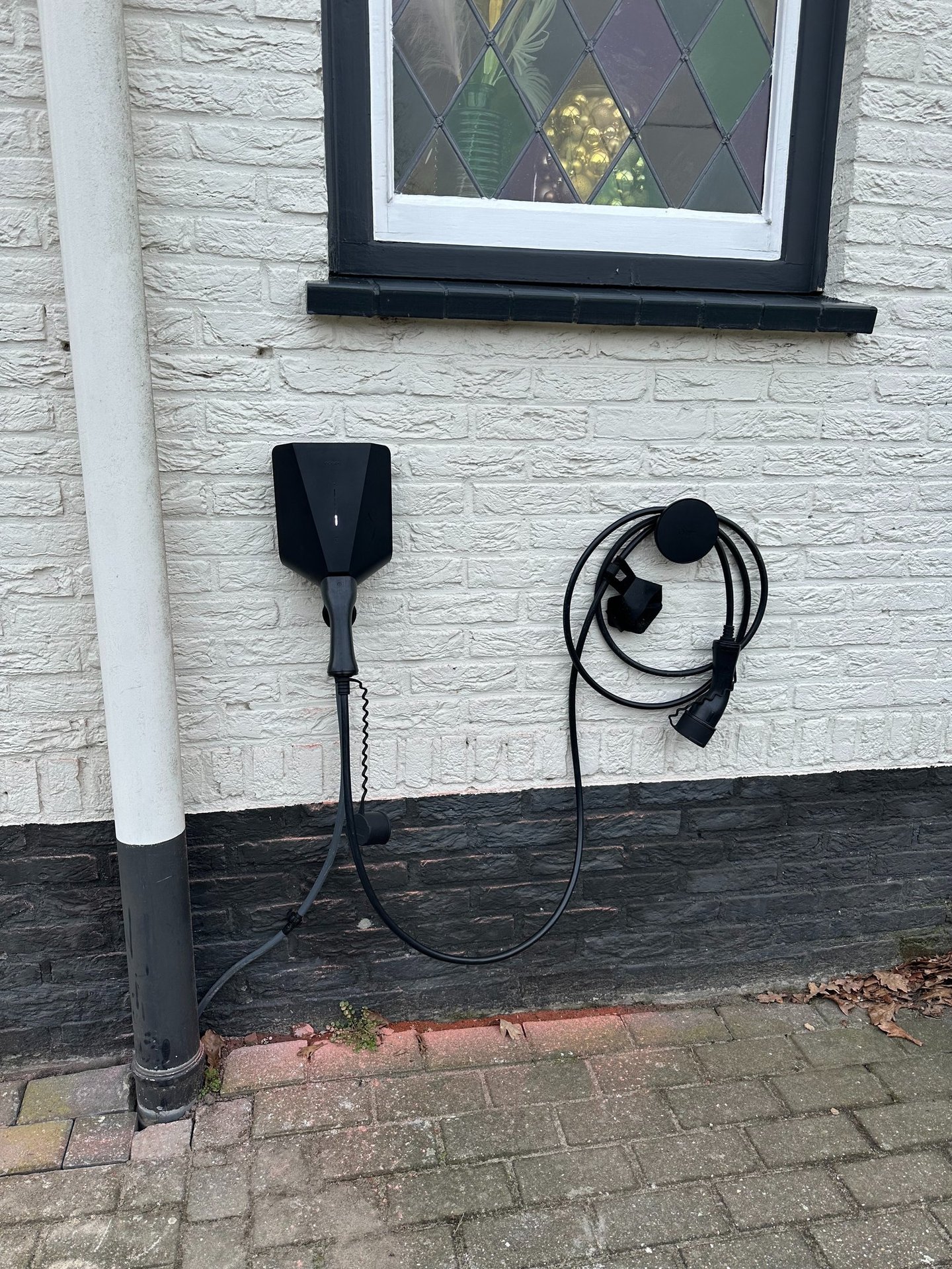 Black wall-mounted electric vehicle charging station and cable installed on a white brick house exterior.