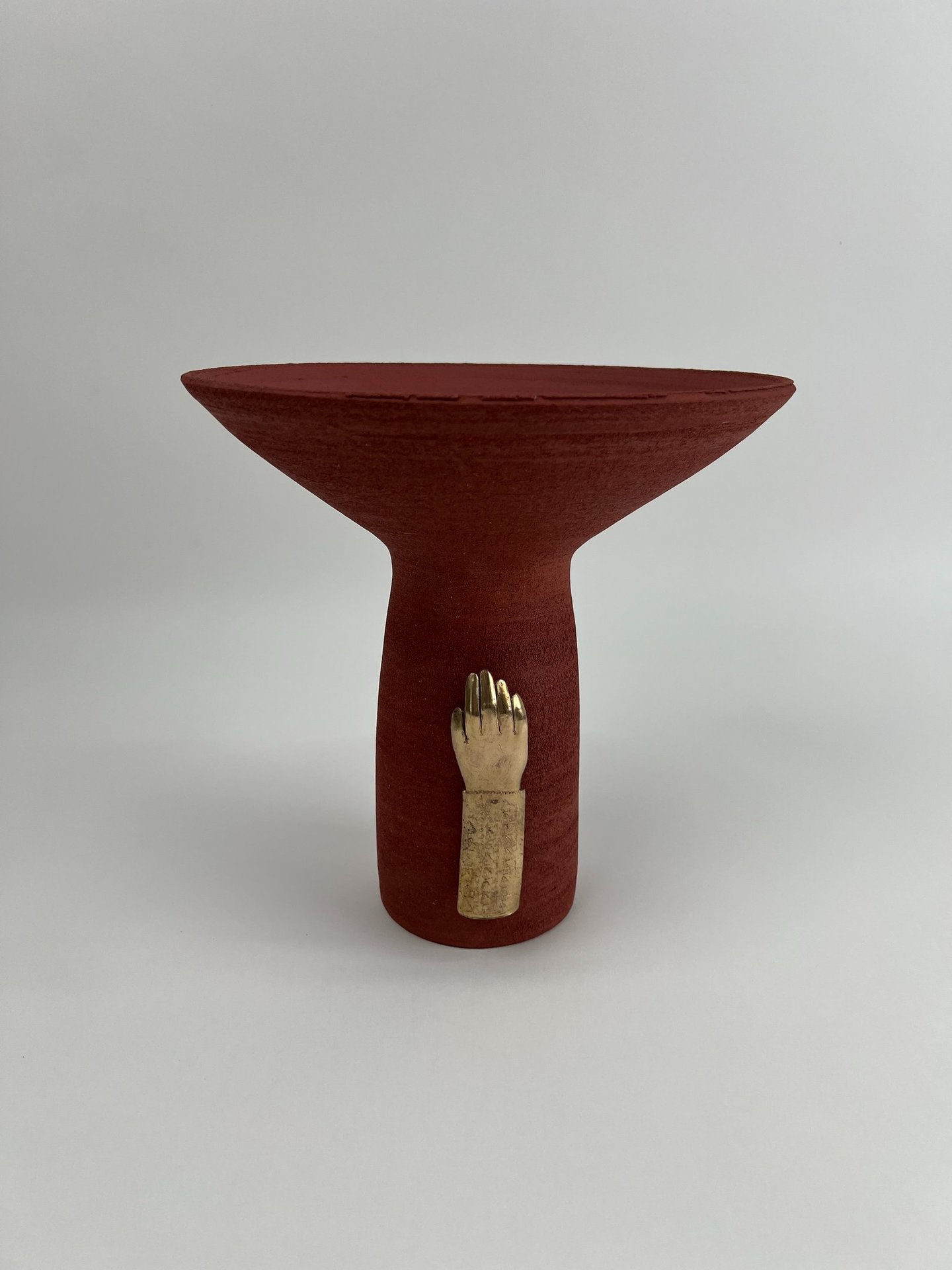 A tall thrown red ceramic vessel, wide at the top. A cast bronze hand is fixed on its base.