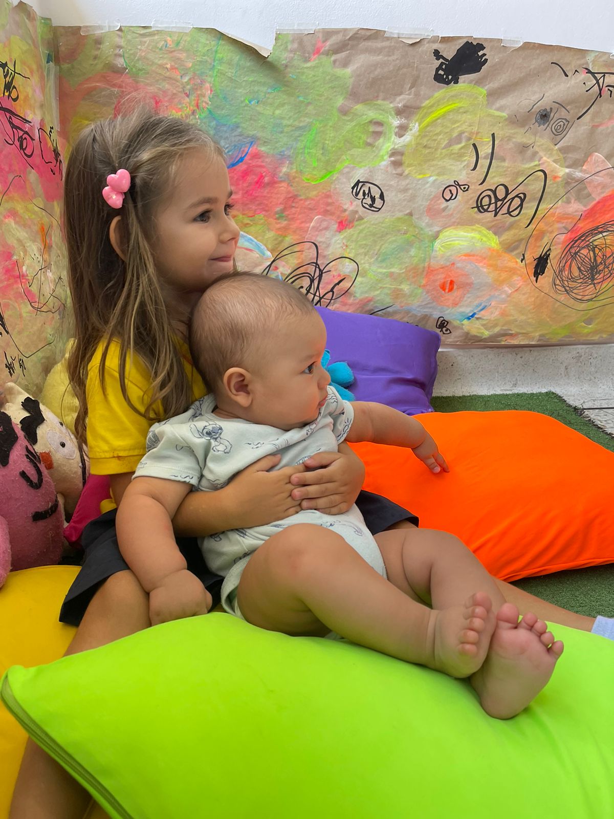Niña abrazando bebe sentados entre cojines de colores vibrantes en Ukelele Jardín Infantil