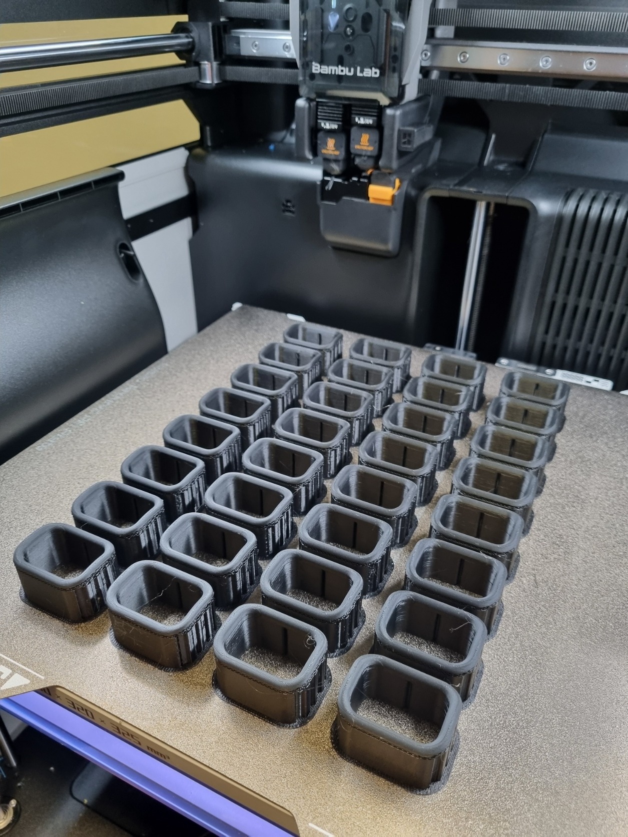 A Bambu Lab 3D printer batch producing black filament components on a textured build plate.