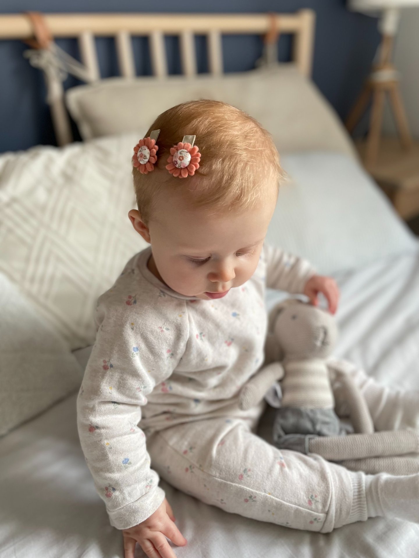 niemowlę siedzi na łóżku z pluszową zabawką, na głowie ma dwie spinki w kształcie kwiatów