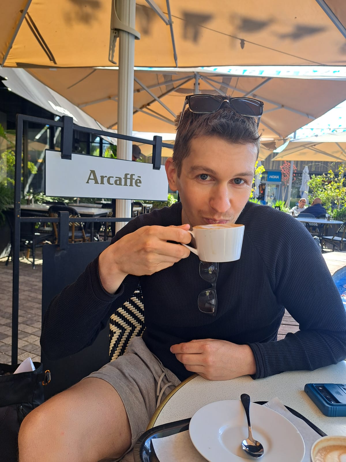 A man in a black long-sleeved shirt drinking a cappuccino at the Arcaffe outdoor cafe terrace.