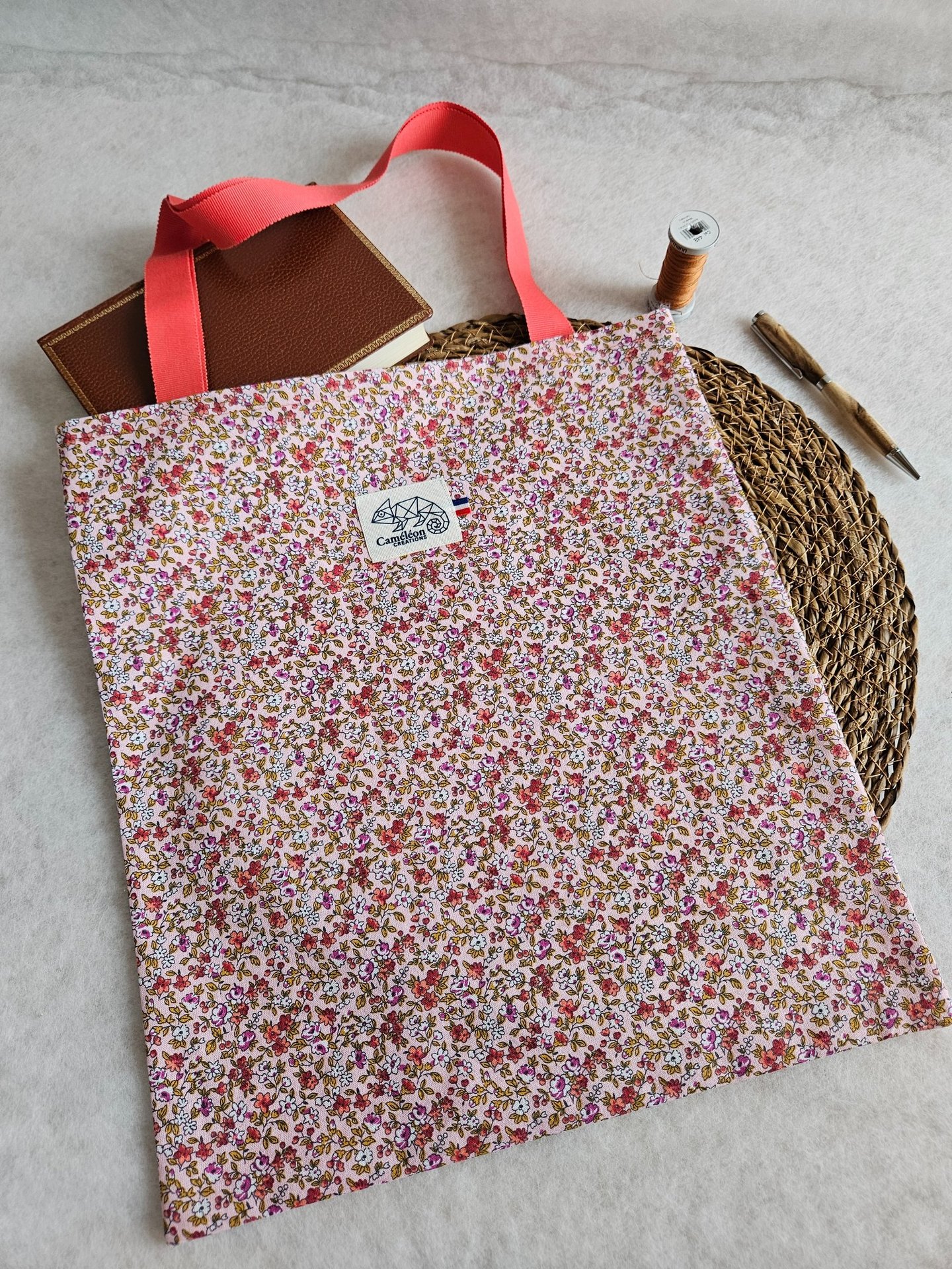 Handmade floral cotton tote bag with pink handles next to a journal, thread, and pen.