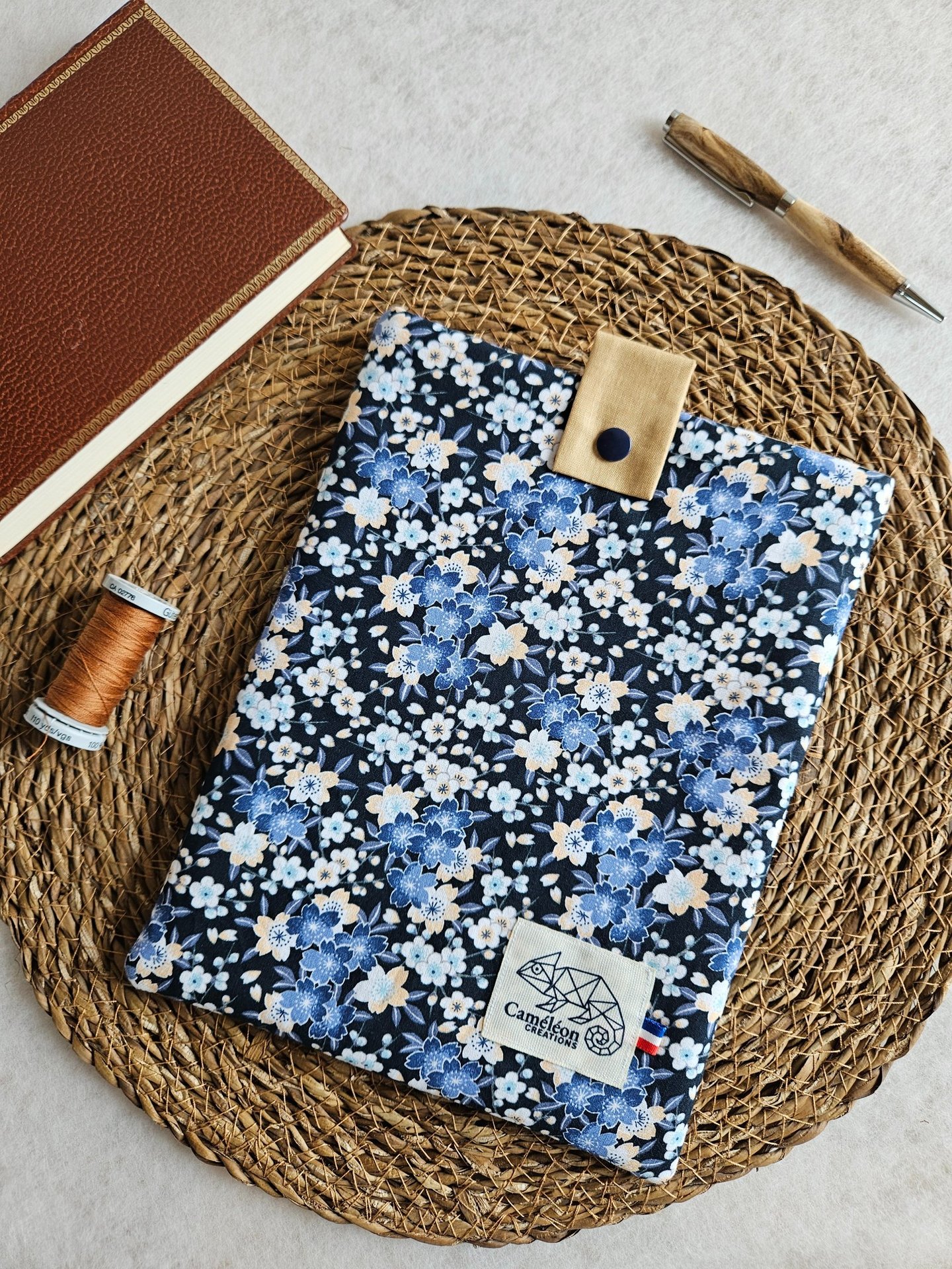 Handmade floral book sleeve with blue cherry blossom pattern, wooden pen, and leather journal on a woven mat.