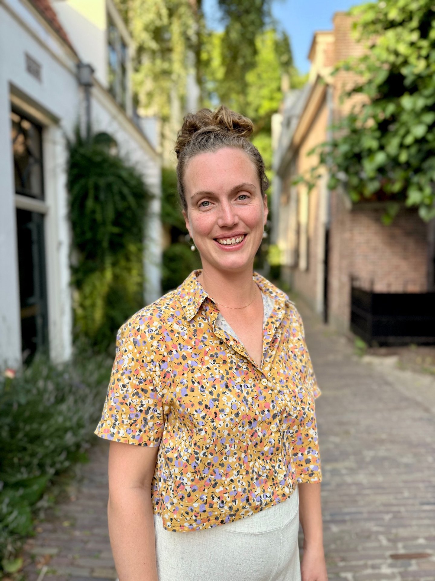 Smiling woman with a hair bun wearing a floral patterned yellow shirt on a cobblestone street.