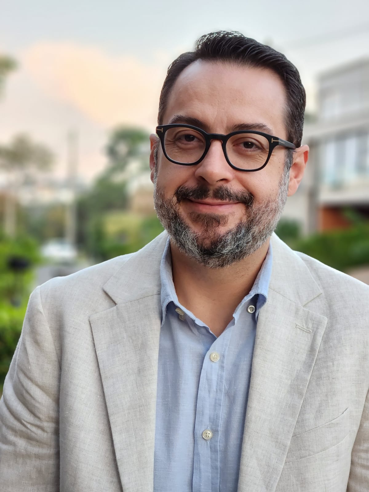 Smiling professional man wearing glasses and a tan blazer against a blurred outdoor background.
