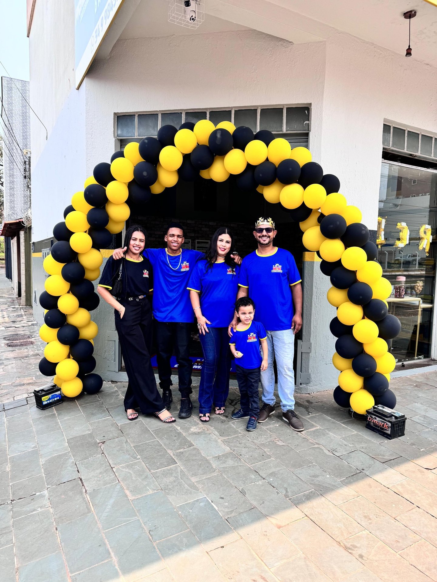 4 adultos e 1 criança com uniformes azuis, em frente a loja de baterias automotivas