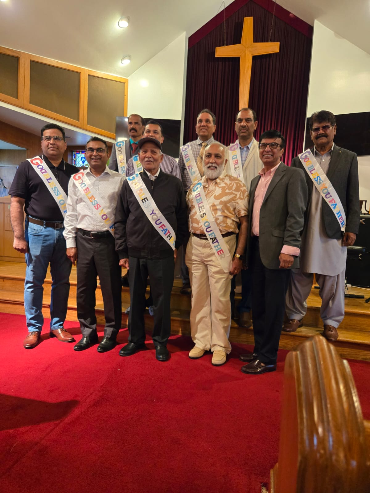 Nine South Asian fathers and father-figures wearing celebratory sashes, standing and smiling under a