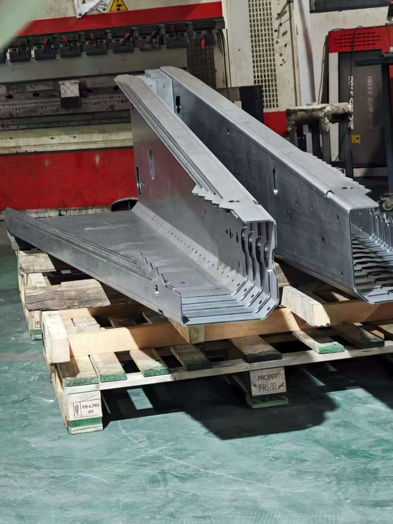 Precision bent galvanized steel sheet metal parts stacked on a wooden pallet in a workshop.