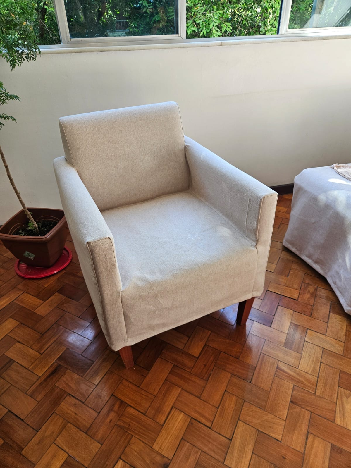 Poltrona estofada bege com pernas de madeira em um piso de parquet em uma sala de estar iluminada.
