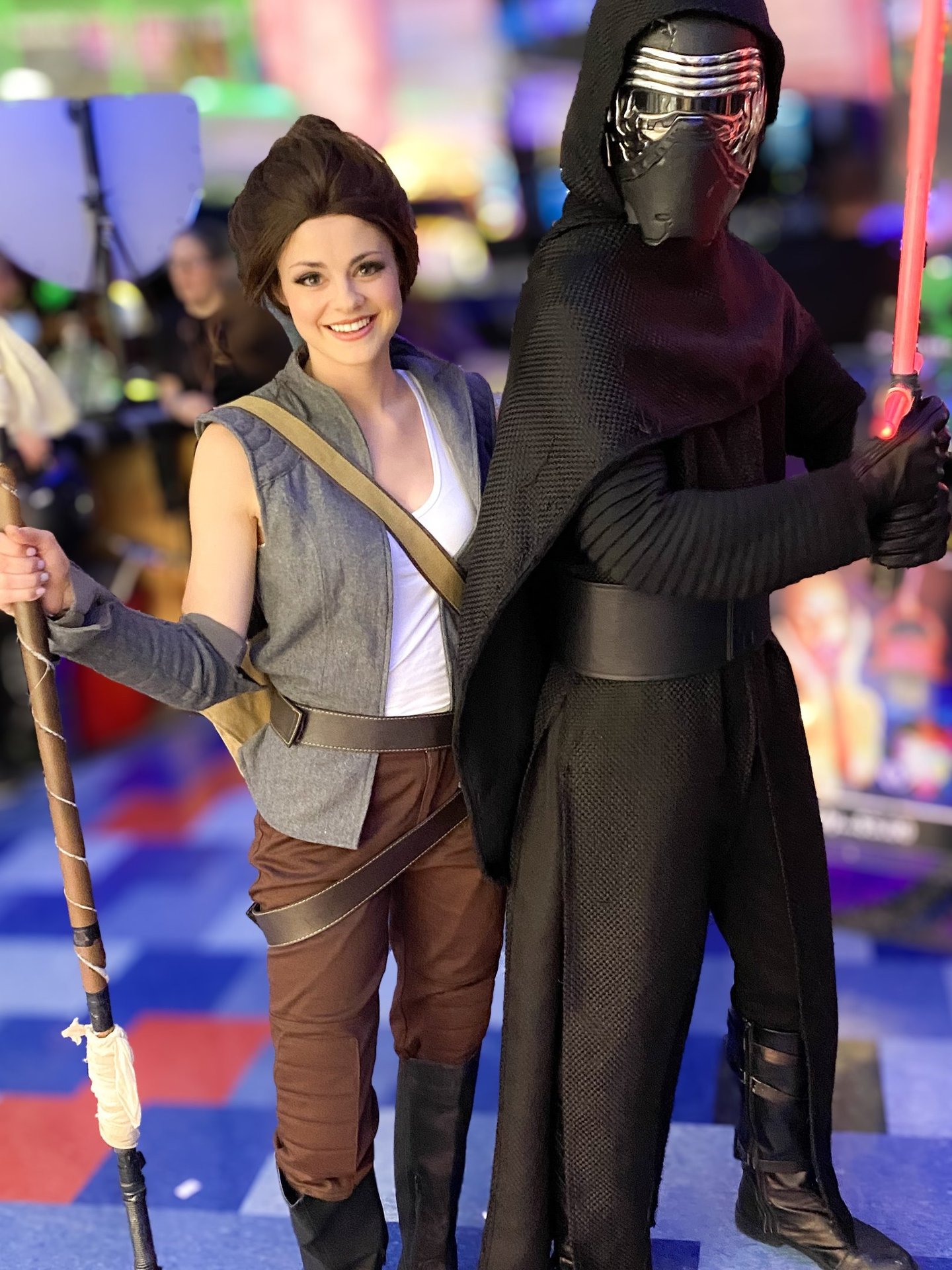 Star Wars fans in Rey and Kylo Ren cosplay posing with a lightsaber and staff at a convention.