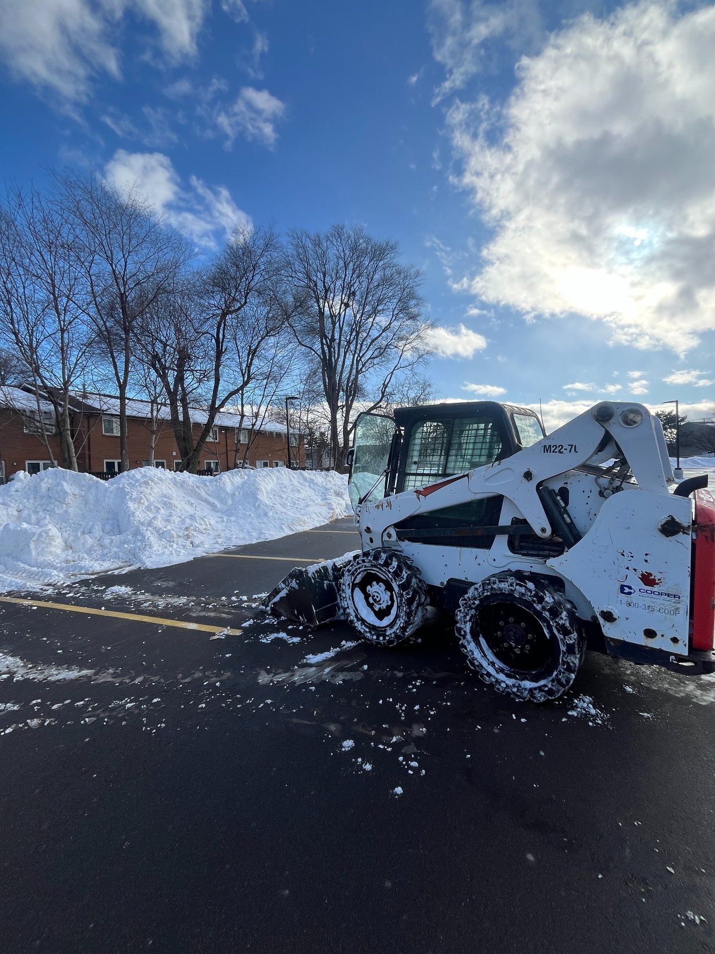 White Bobcat skid steer loader clearing a snow-covered commercial lot near residential buildings.