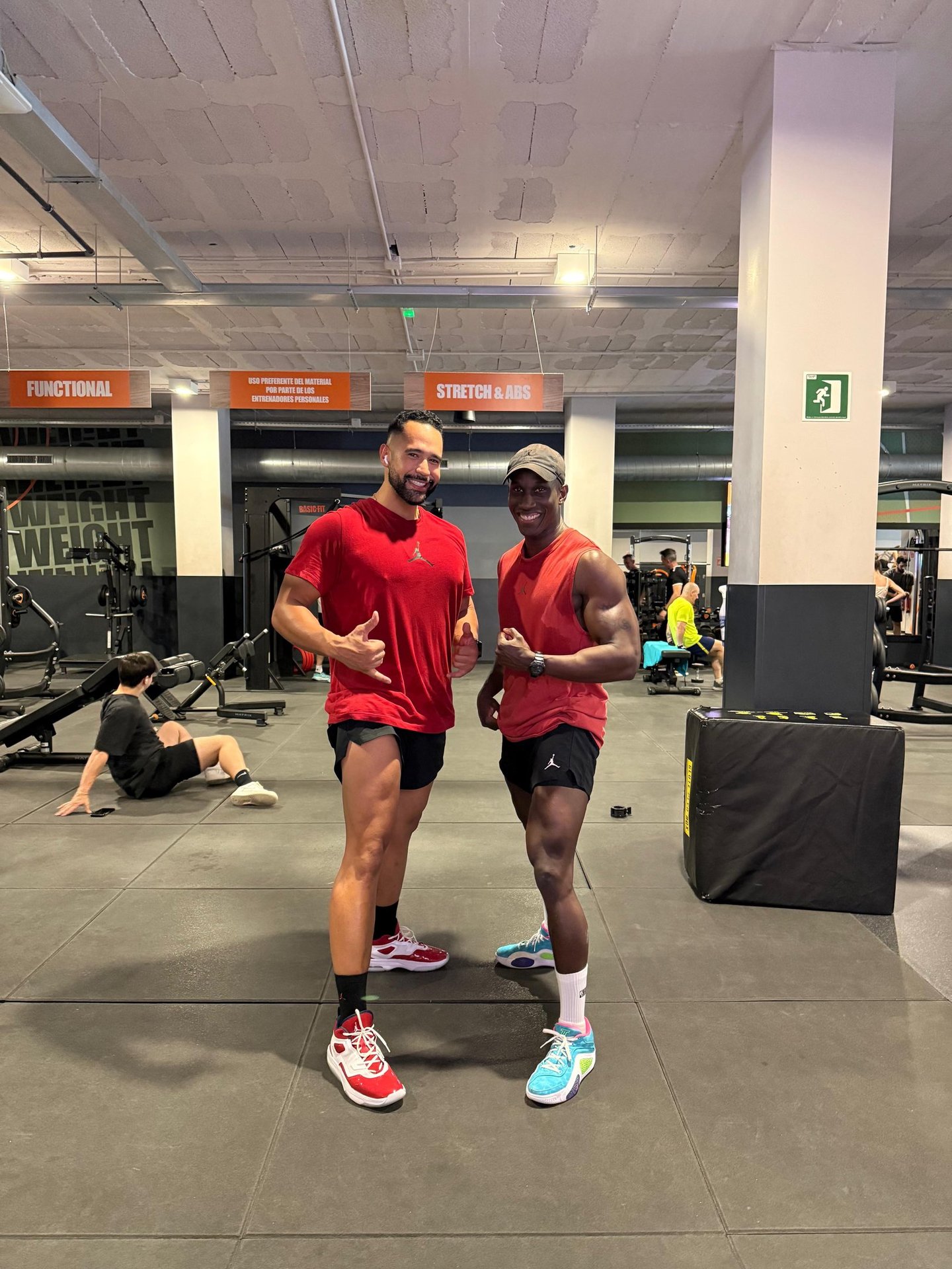 Two muscular men smiling and posing for a fitness photo inside a modern gym with exercise equipment.
