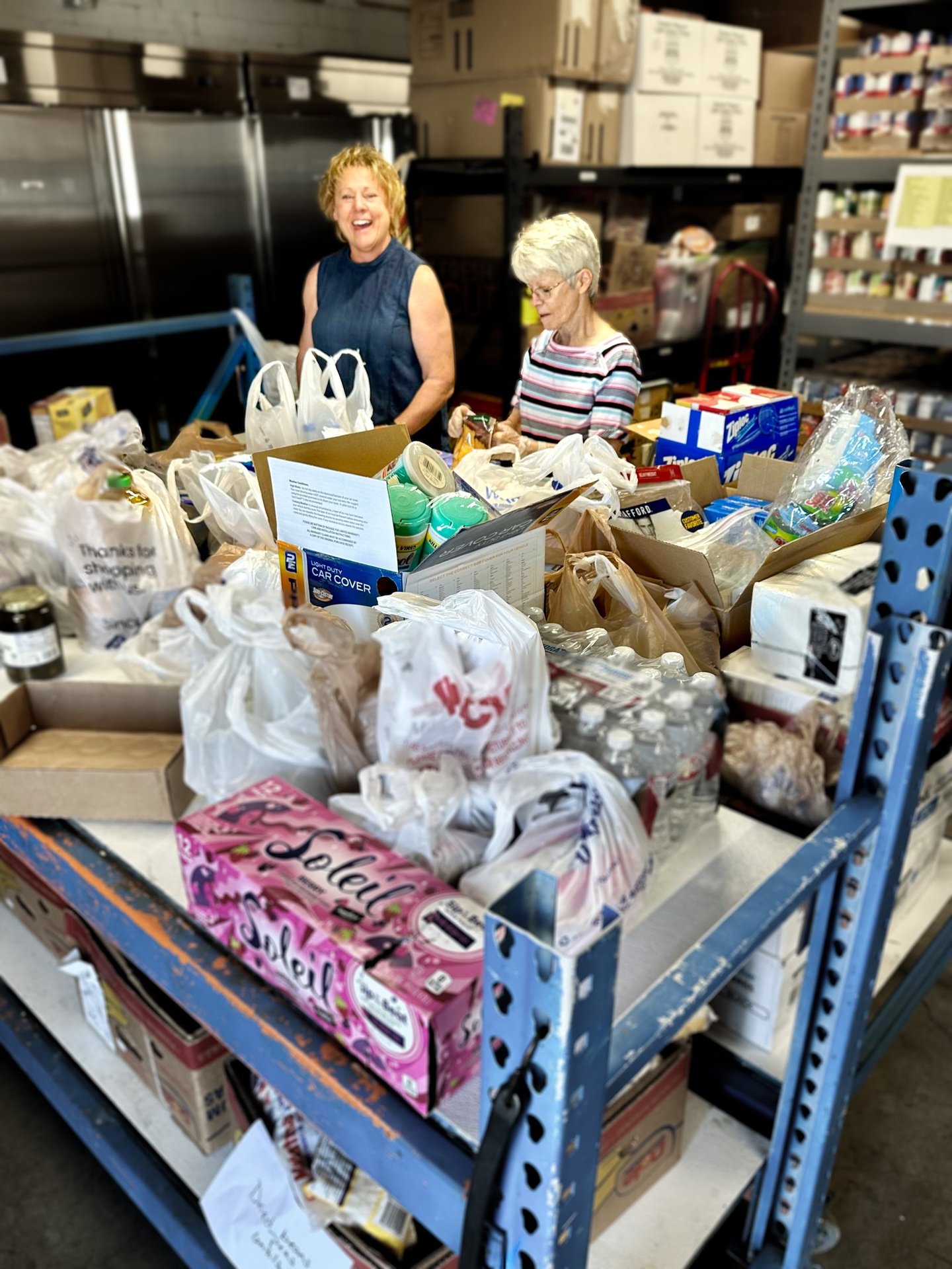 Two women standing behind table covered in bags of donated food in food pantry 