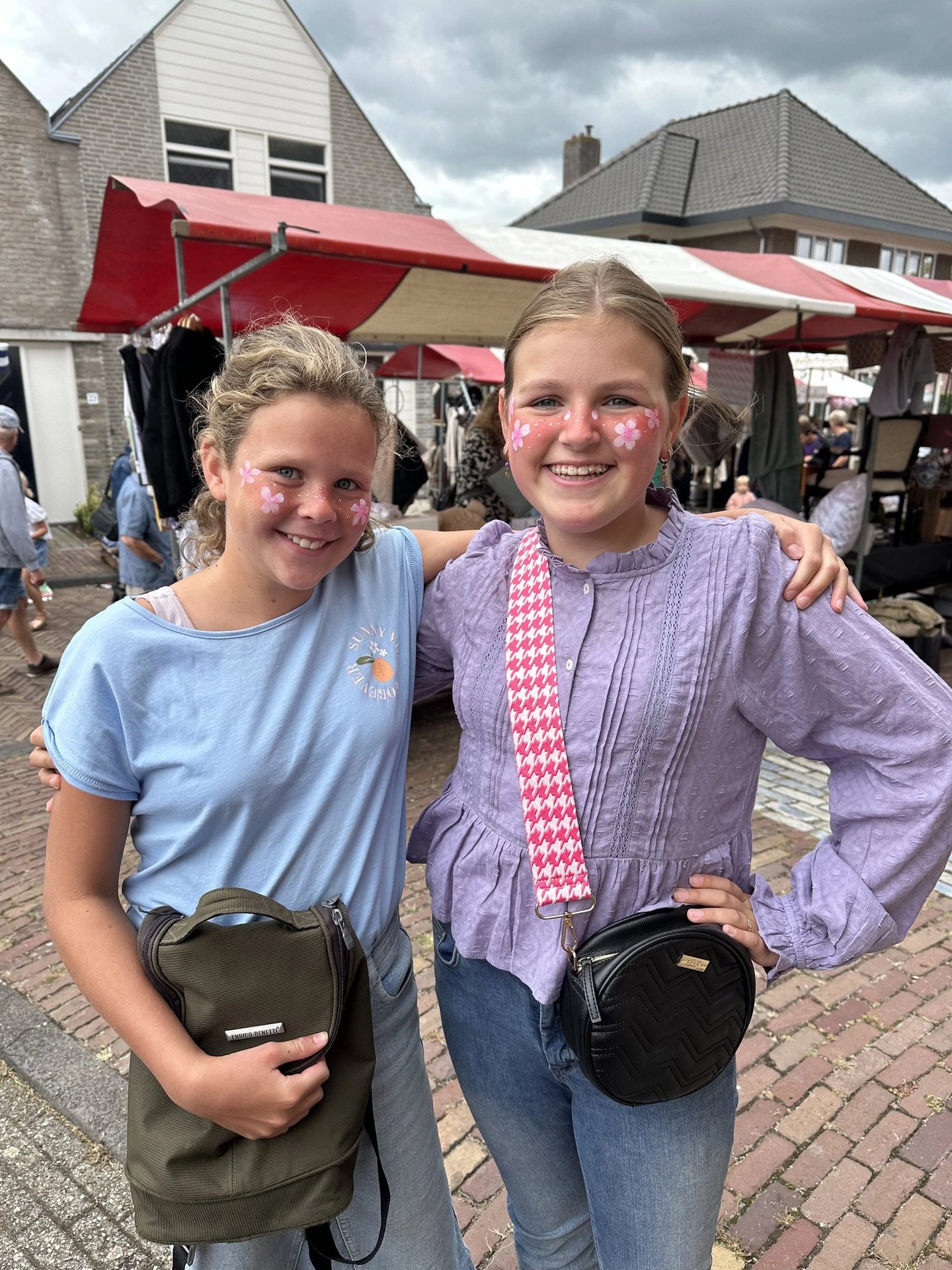 Twee lachende meiden met matchende roze bloemensmink op hun wangen poseren op een markt.