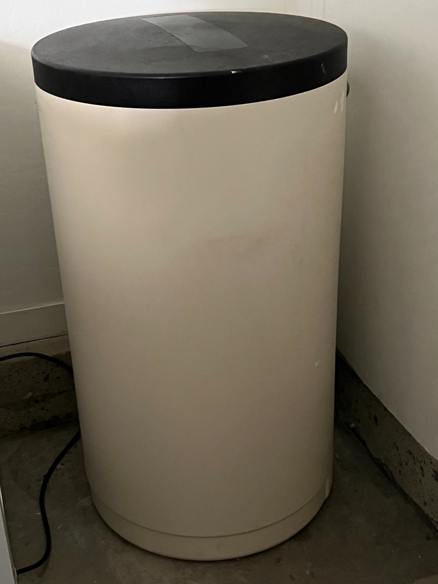 A cylindrical beige water softener brine tank with a black lid in a utility room corner.