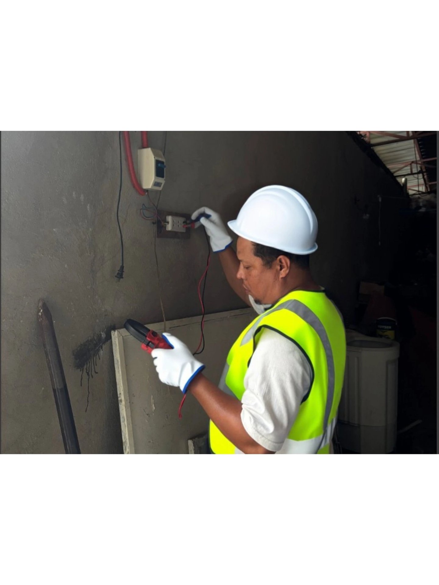 An electrician in a hard hat and safety vest uses a multimeter to test a wall power outlet.