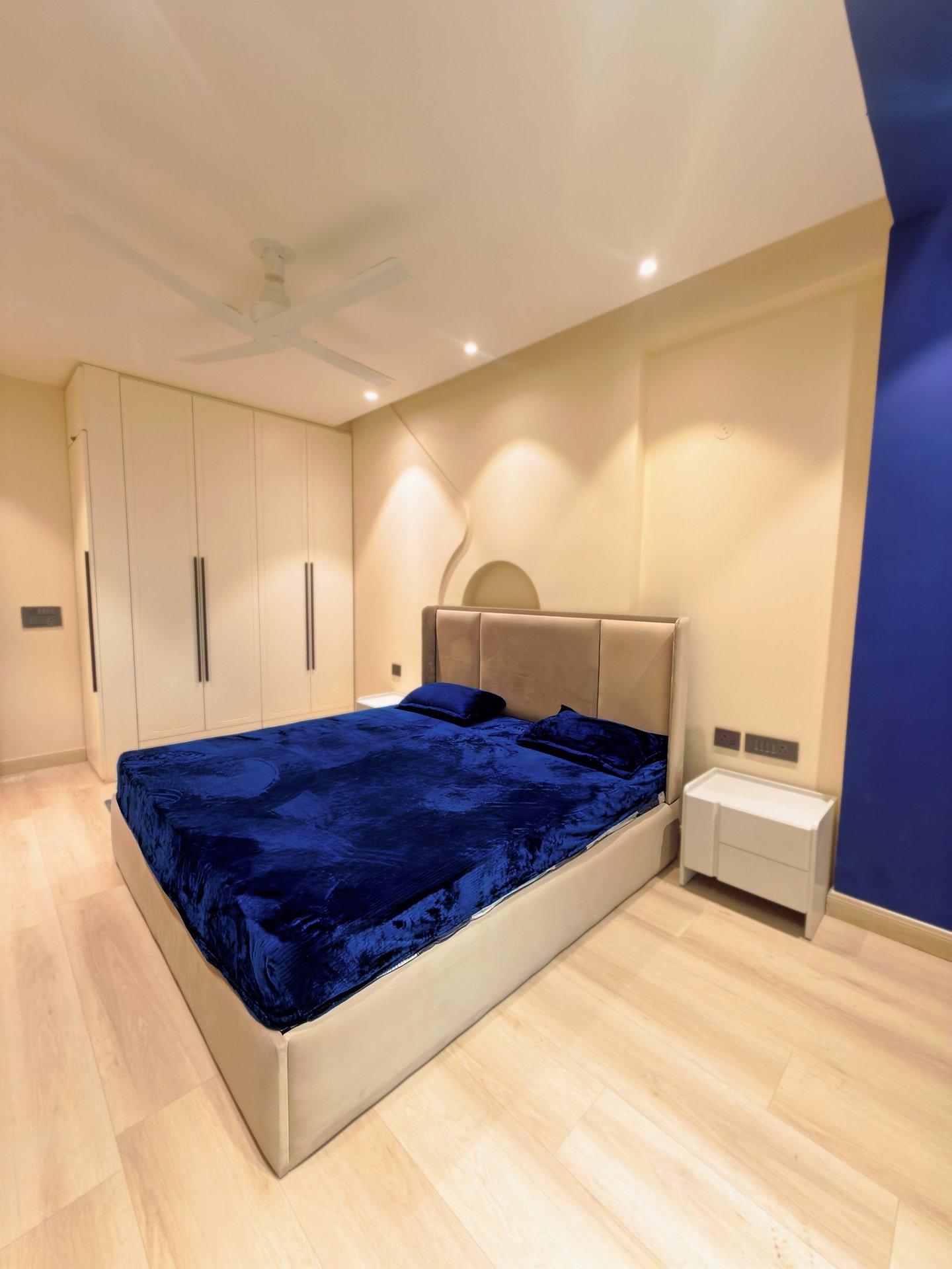 Modern master bedroom with a blue velvet bedspread, white wardrobe, and recessed lighting.