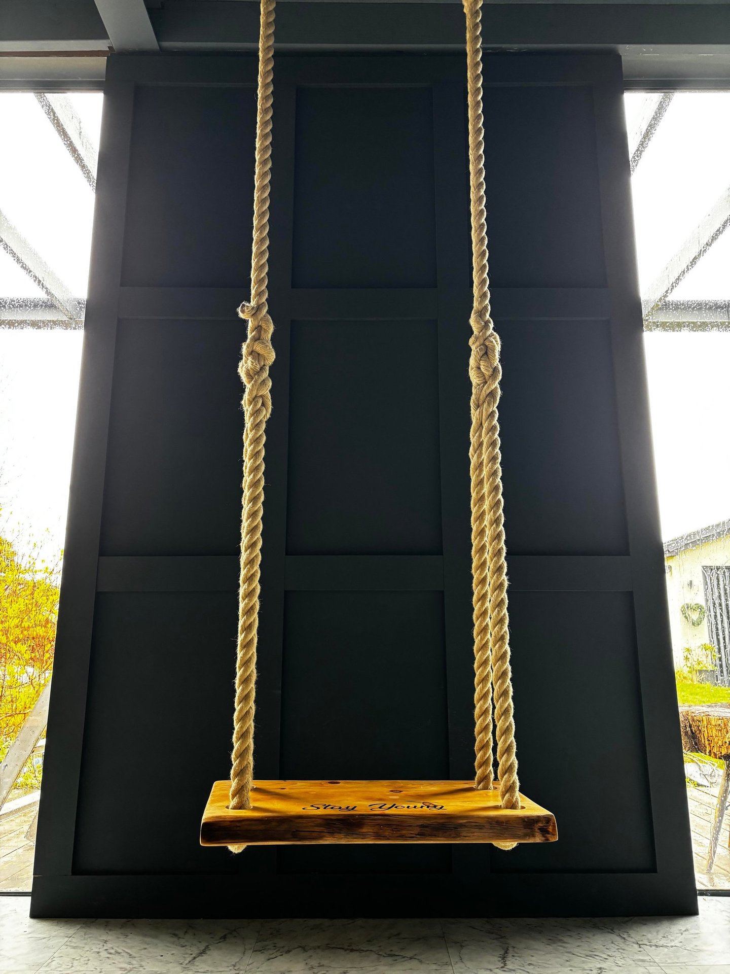 Indoor wooden swing with thick ropes hanging against a black paneled accent wall.