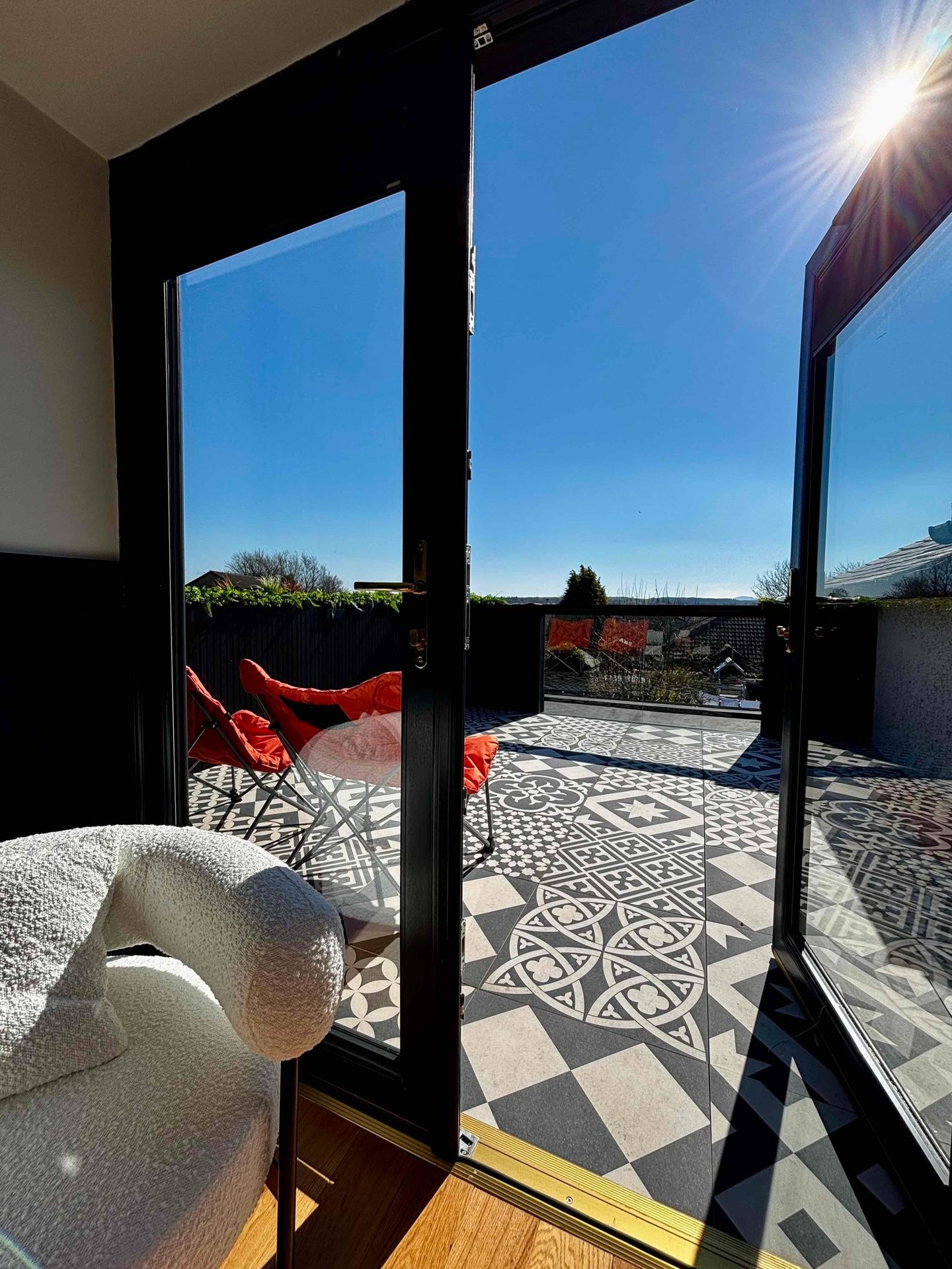 Modern rooftop terrace with patterned tile flooring, red chairs, and black French doors under a sunny blue sky.