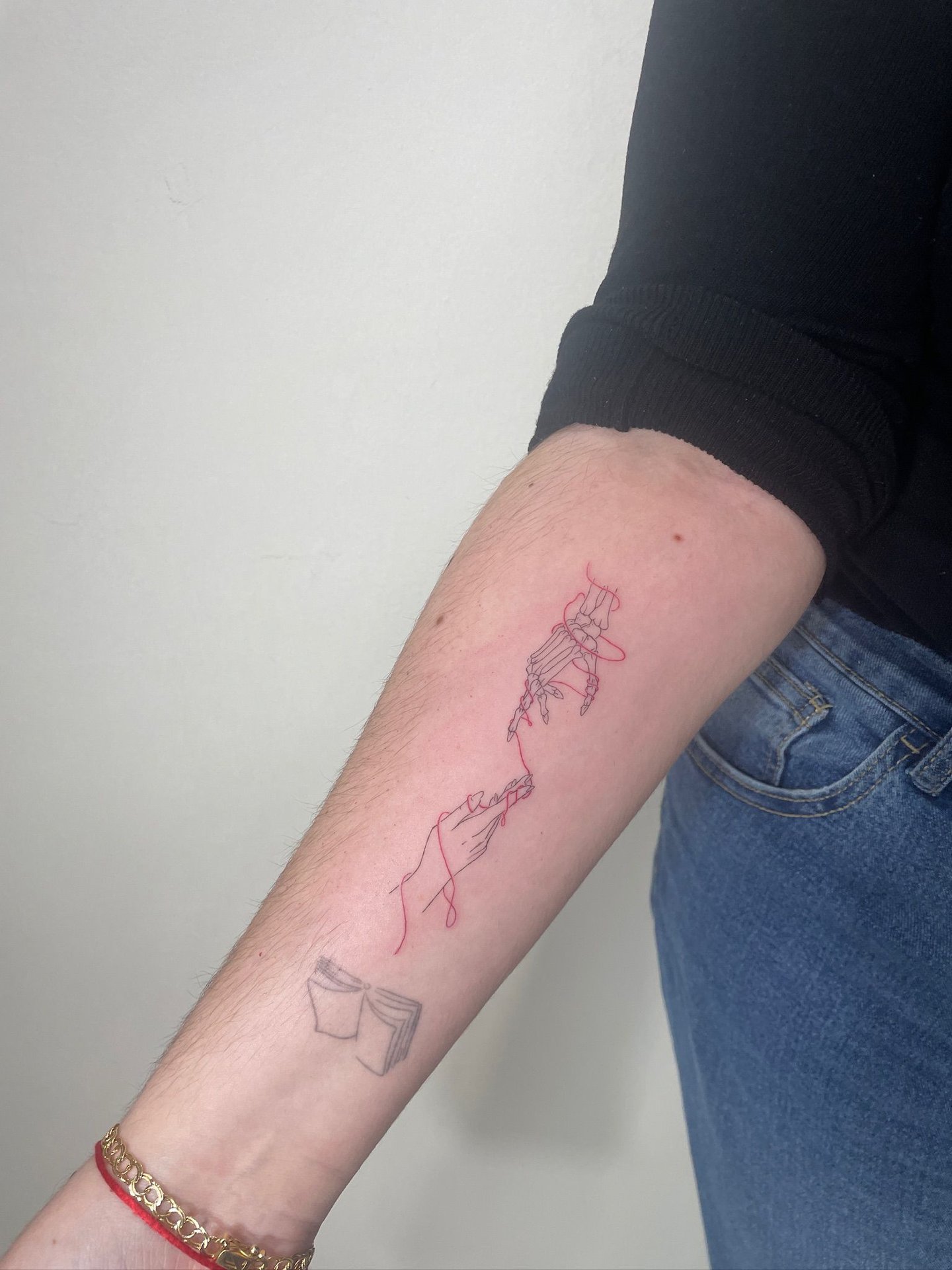 Tatuaje fineline en Madrid de mano esqueleto y mano humana unidas por hilo rojo con libro abierto