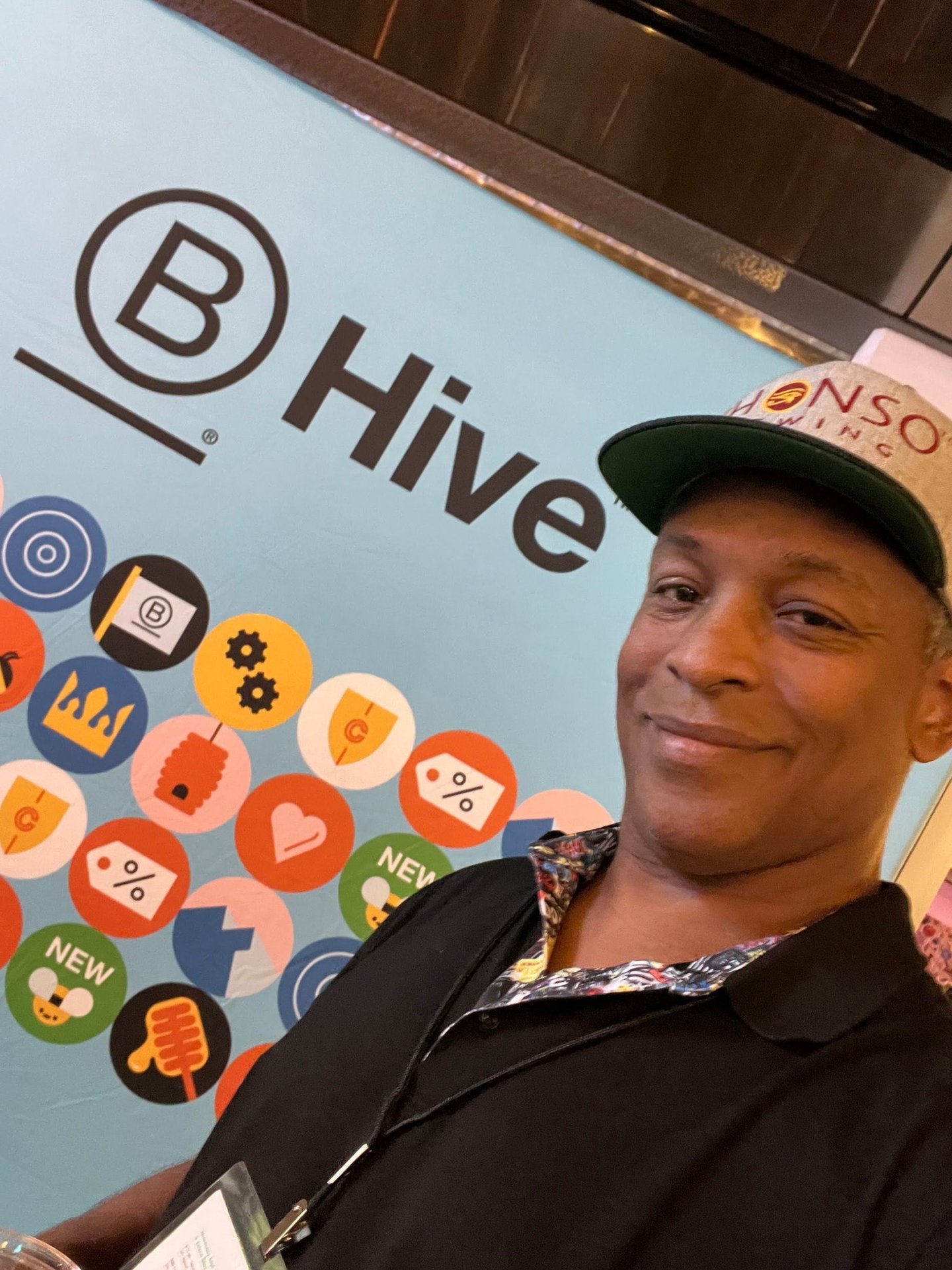A man in a baseball cap smiling in front of a blue B Hive brand wall with colorful icons.