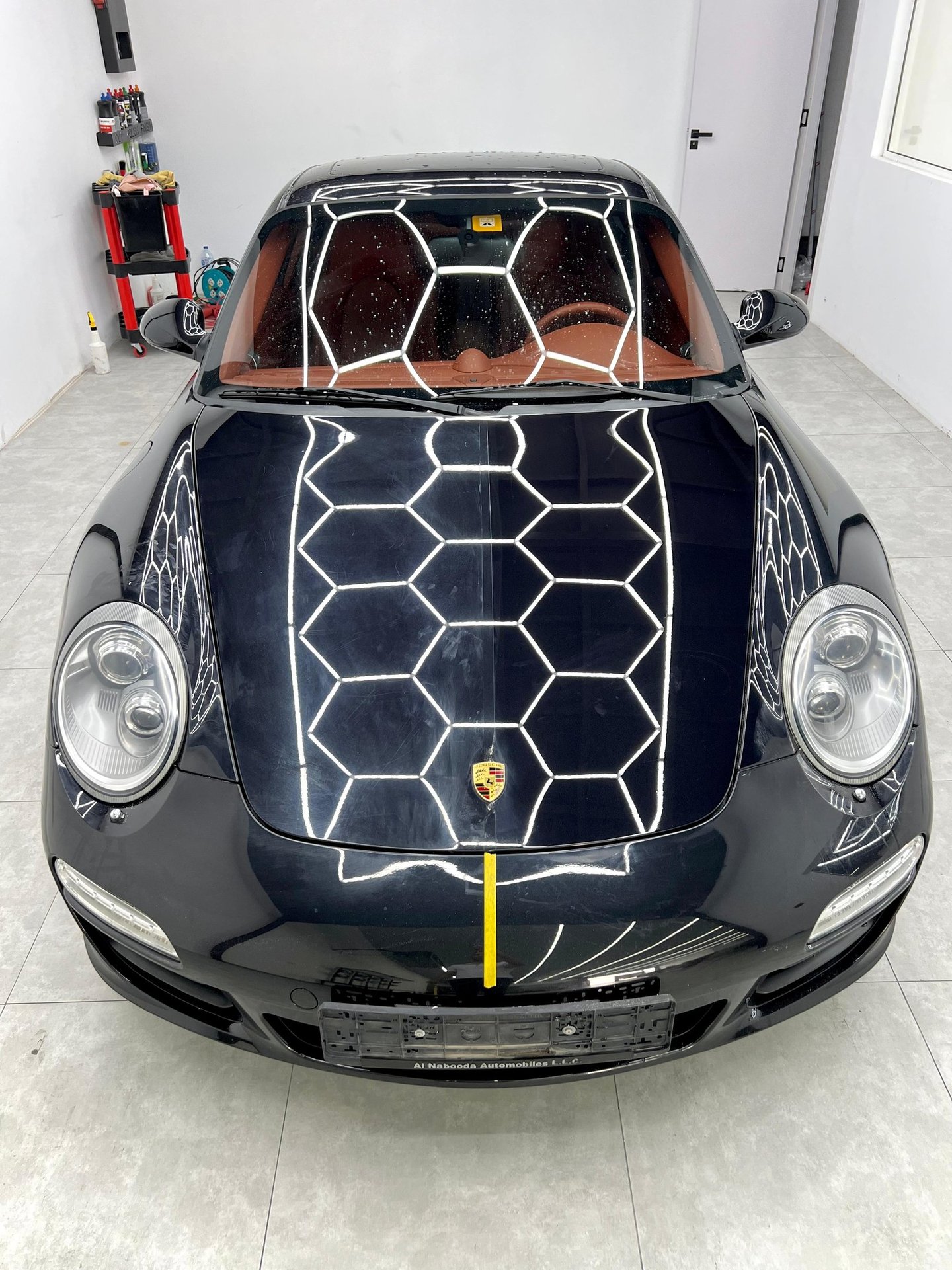 Black Porsche 911 luxury sports car with reflective honeycomb ceiling lights in a professional detailing studio.