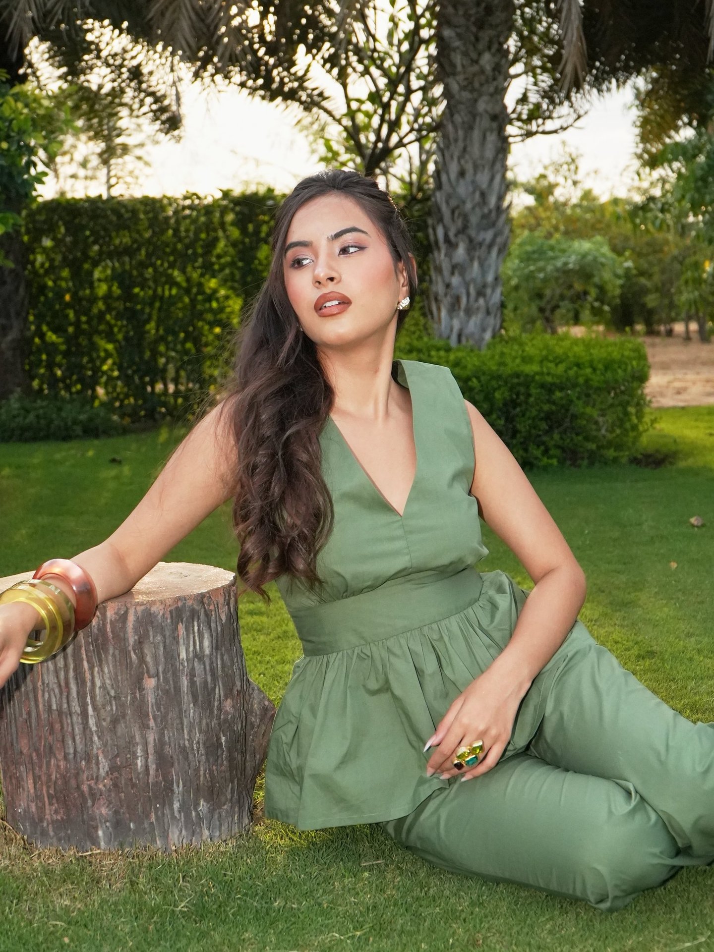 A woman in a sage green peplum jumpsuit sitting on a garden lawn leaning against a wooden tree stump.