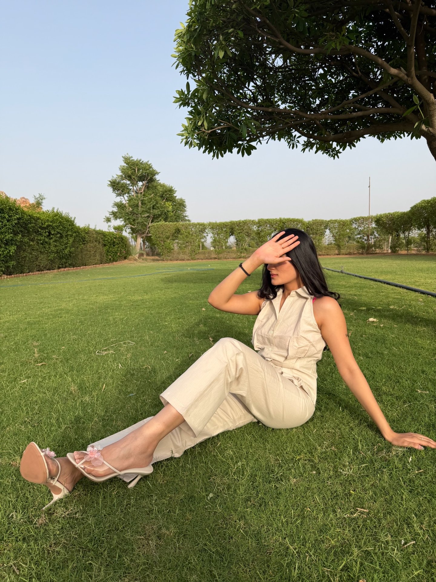 Woman in a beige jumpsuit and high heels sits on a green lawn under a tree, posing with hand over eyes.