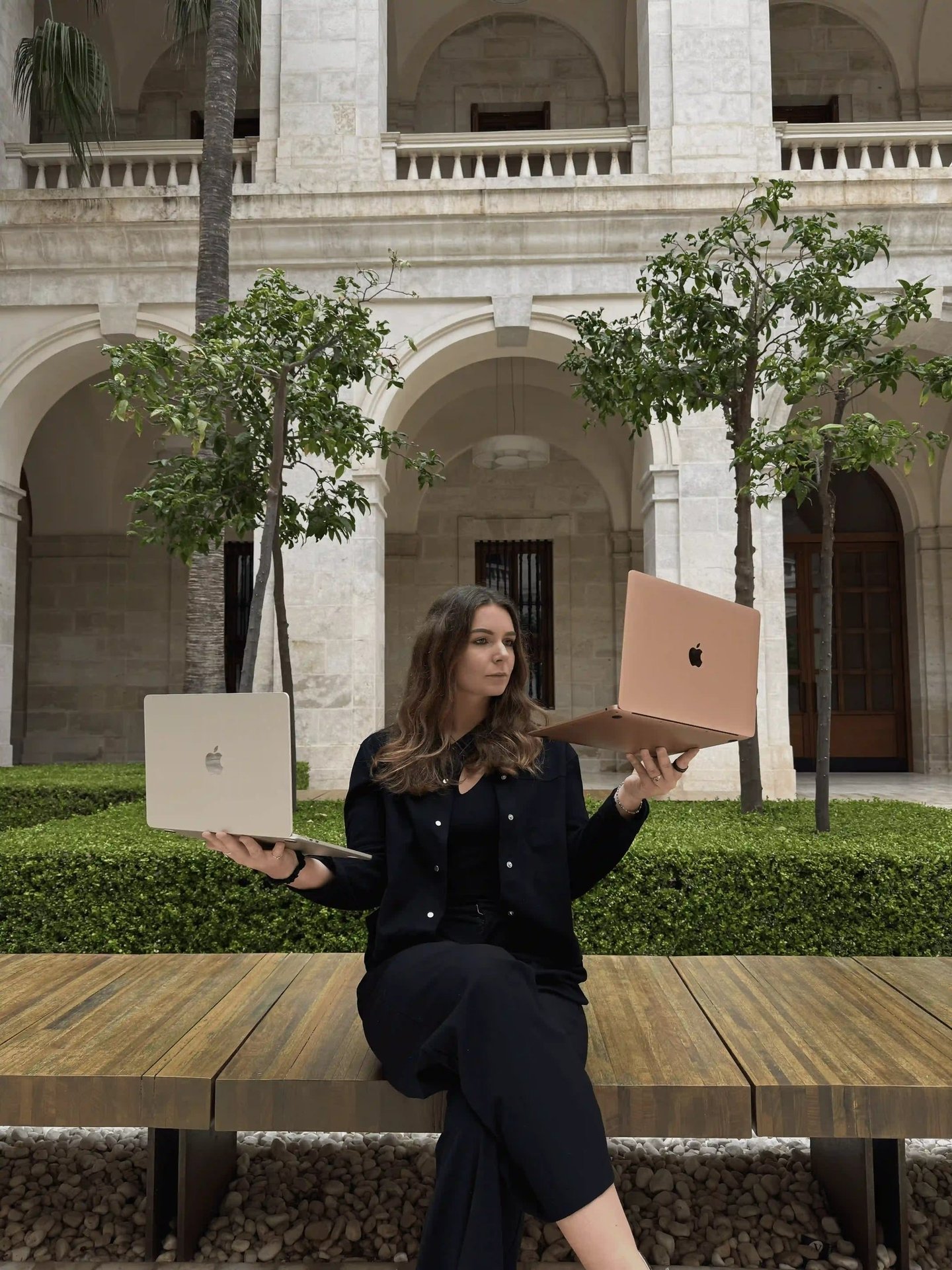 A woman balances two Apple MacBooks while sitting in a stone courtyard, illustrating remote work technology.