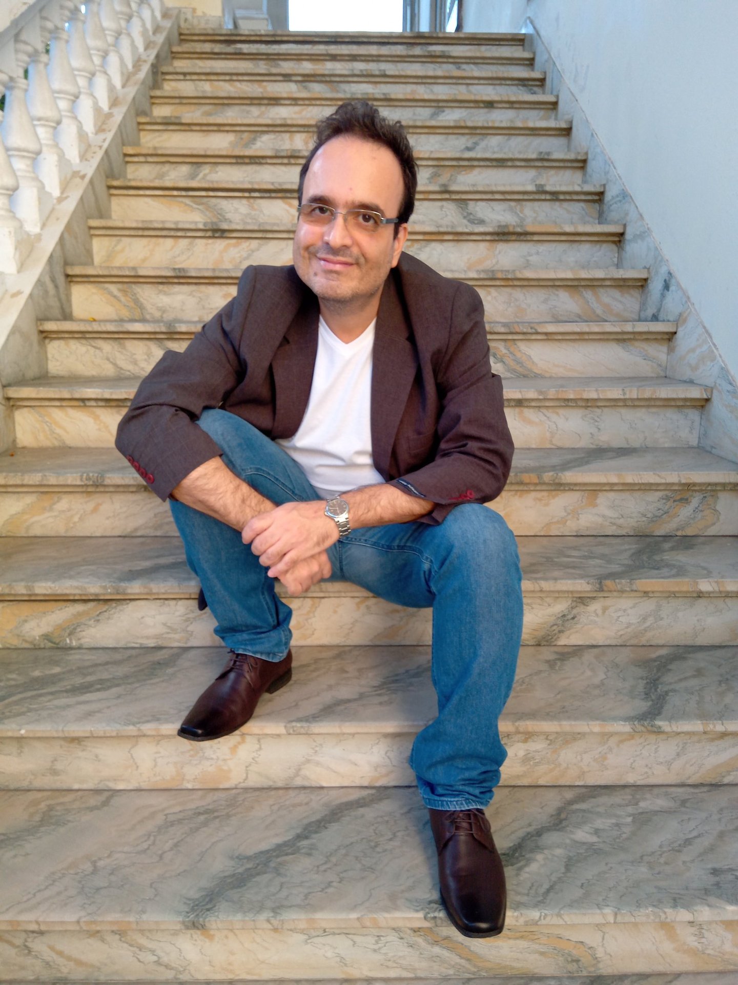 Jornalista Marcelo Dias, sentado à uma escadaria de mármore, de calça jeans, camisa branca e blazer.