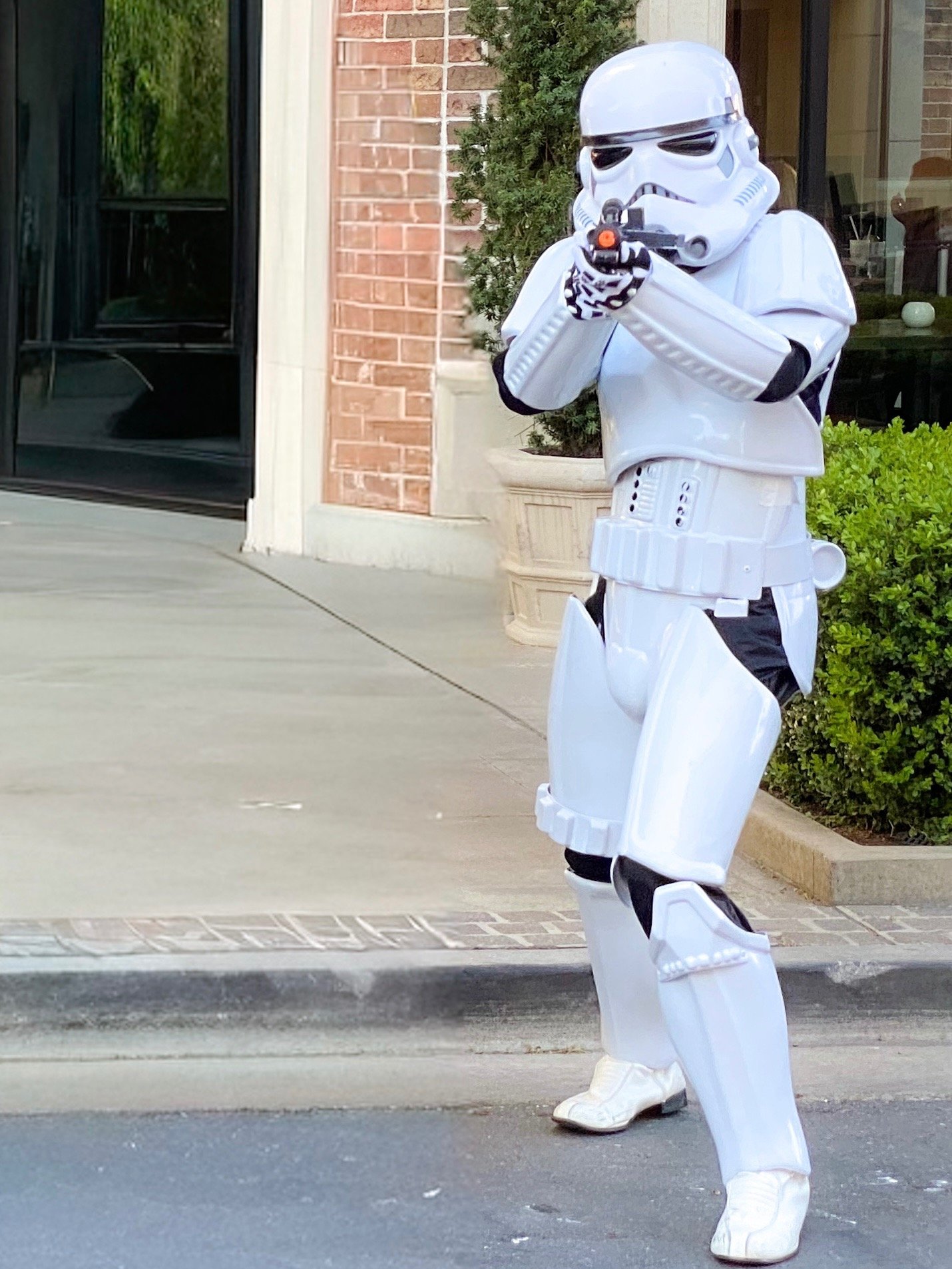 A Star Wars Stormtrooper in full white armor aims a blaster rifle while standing on a sidewalk.
