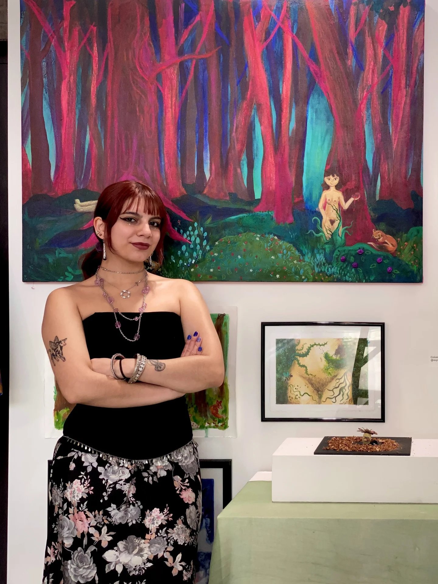 Artist with crossed arms posing in front of her colorful abstract forest paintings in a gallery.