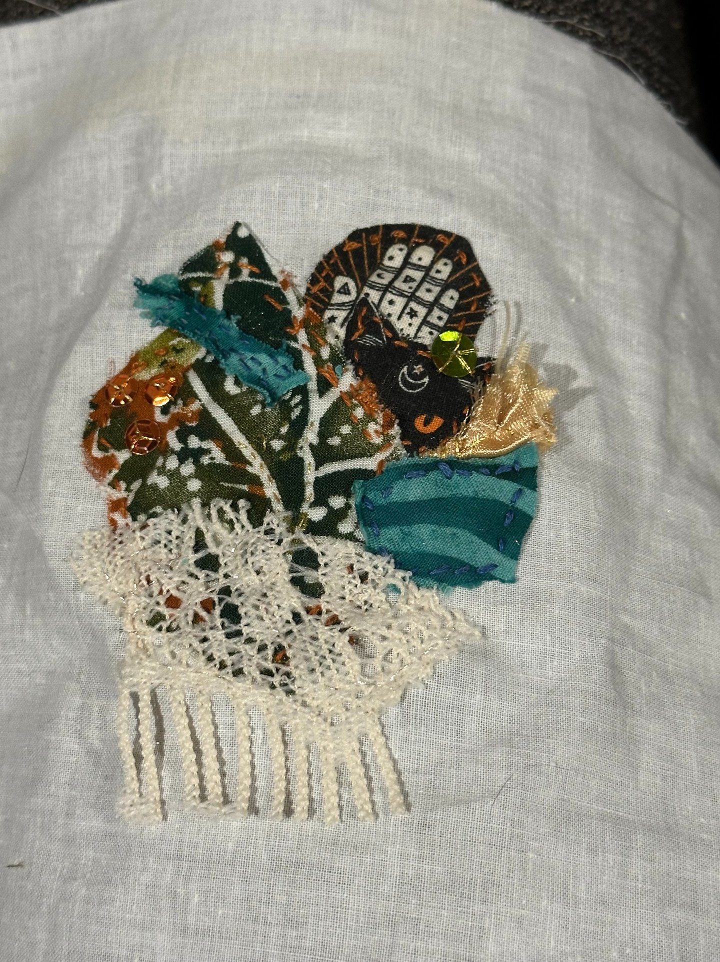 fabric collage with sequins in shades of green, teal, rust, black and cream; cat face and lace used