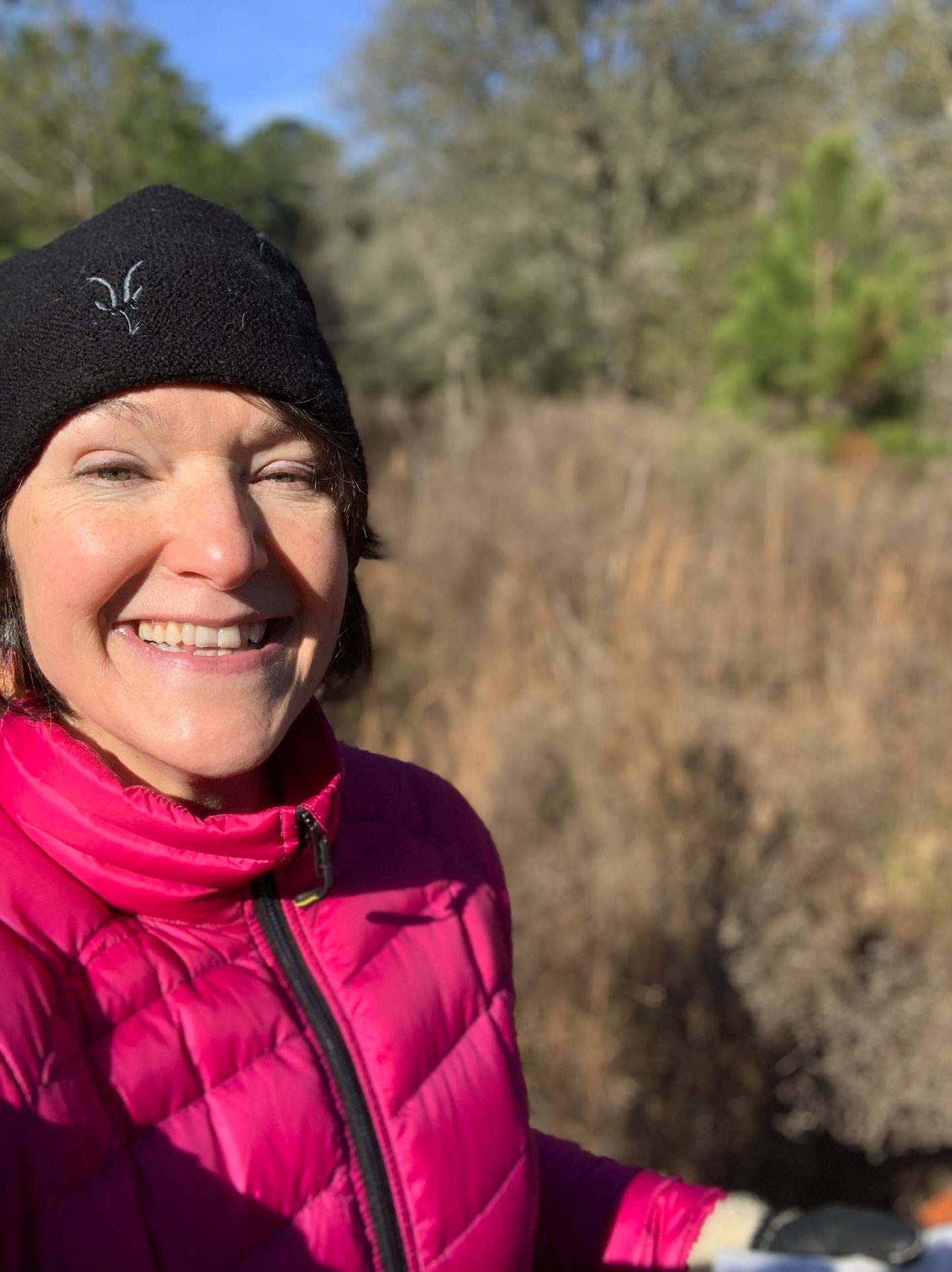 A smiling woman wearing a pink puffer jacket and black beanie hiking outdoors on a sunny day.