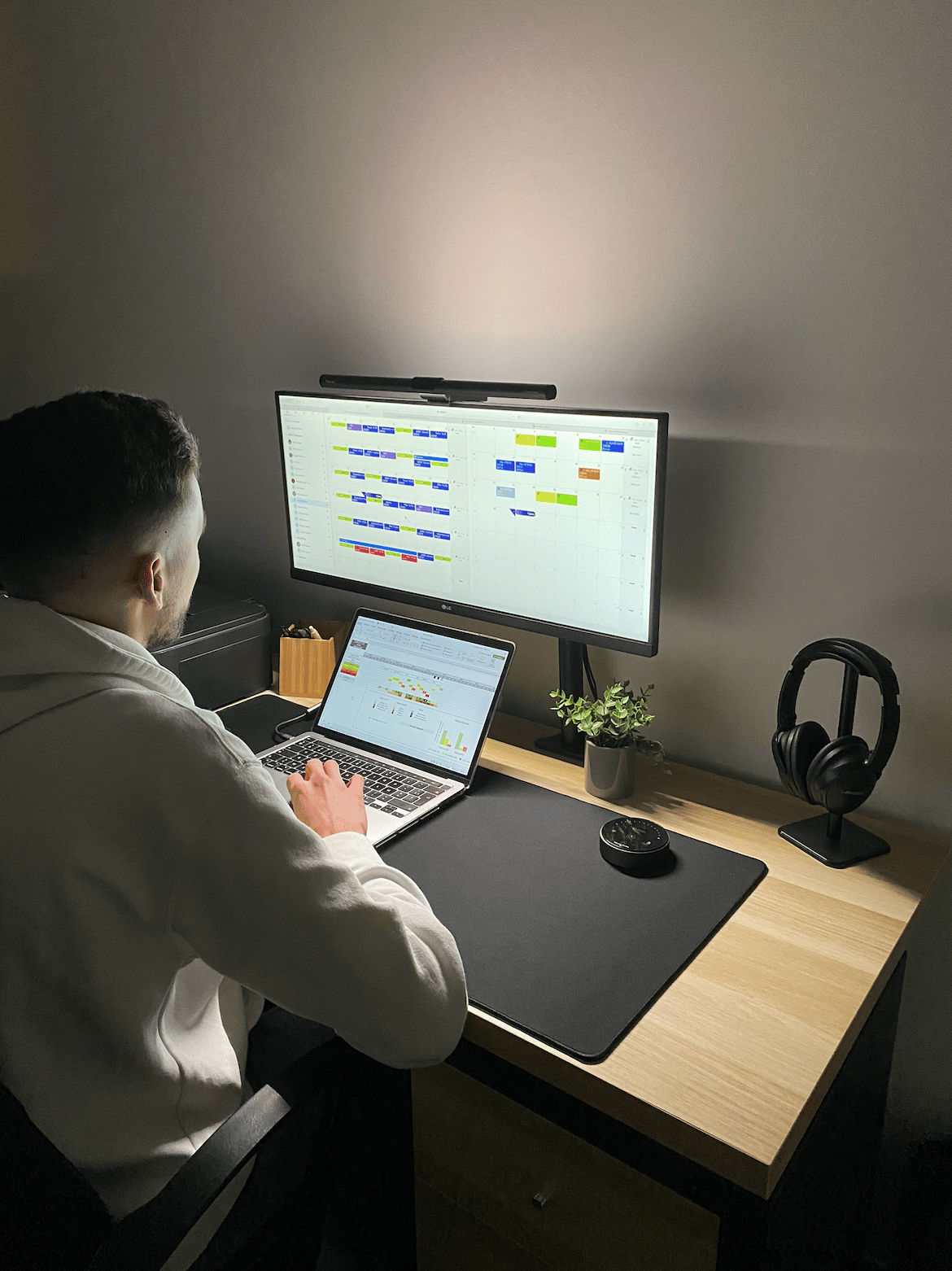 A man working at a home office desk setup with a laptop, monitor, and screen bar light.