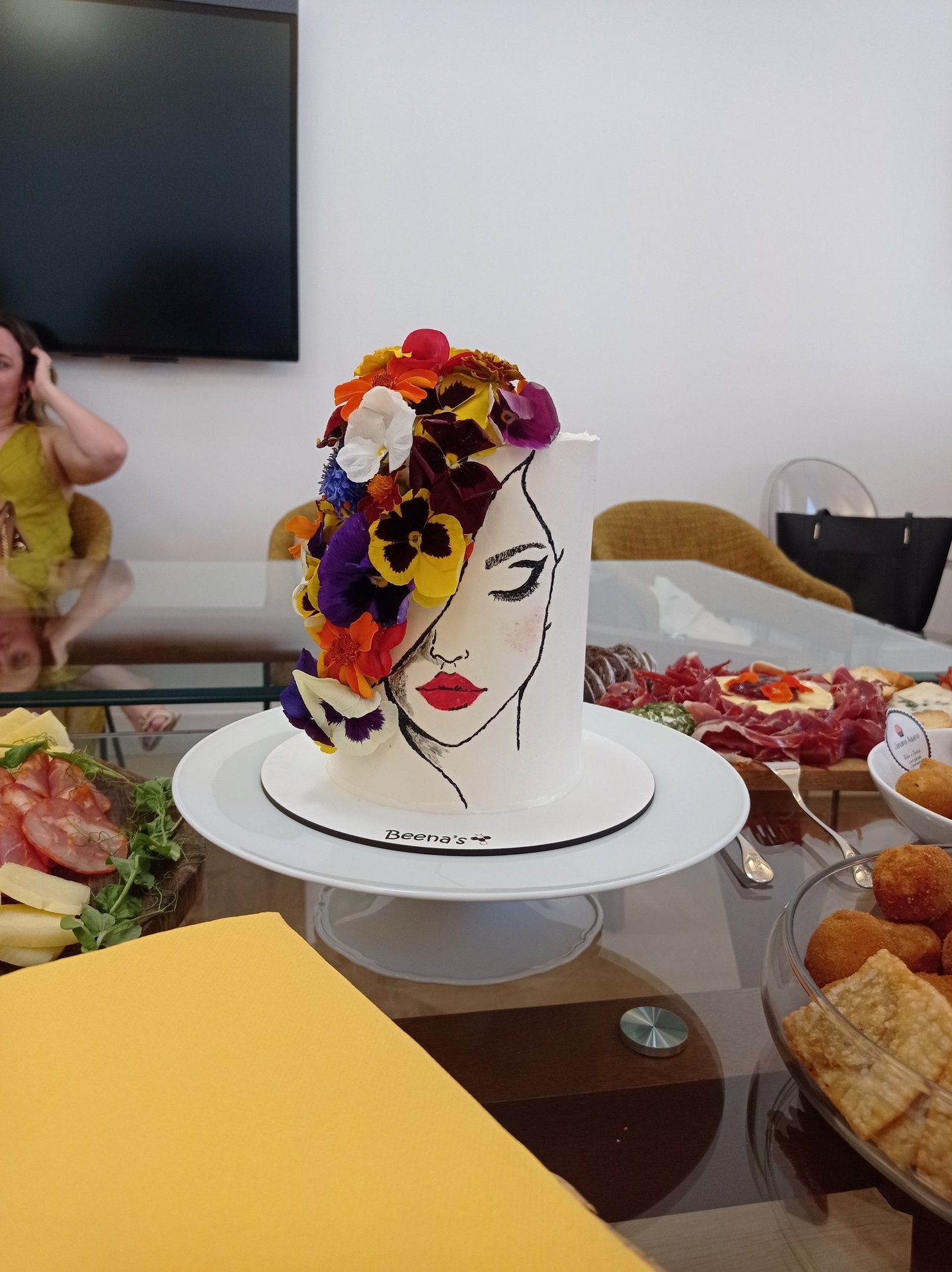 Elegant tall white cake featuring a hand-painted woman's face and colorful edible flowers.