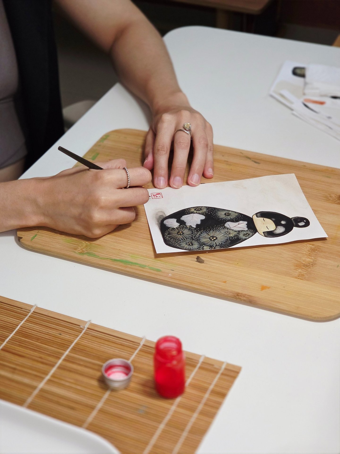 Artist painting a Japanese Kokeshi doll illustration on paper with a fine brush and red ink.