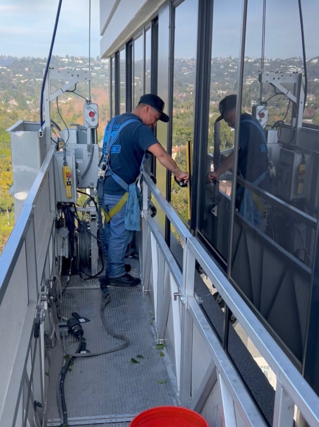 Professional window cleaner in safety harness using a squeegee on high-rise office windows from a platform.